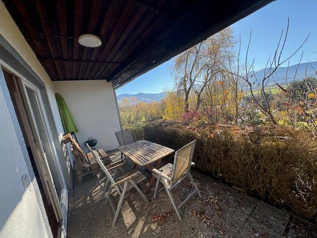 Überdachte Terrasse mit Holztisch und Stühlen, umgeben von Hecken und Blick auf die Landschaft.