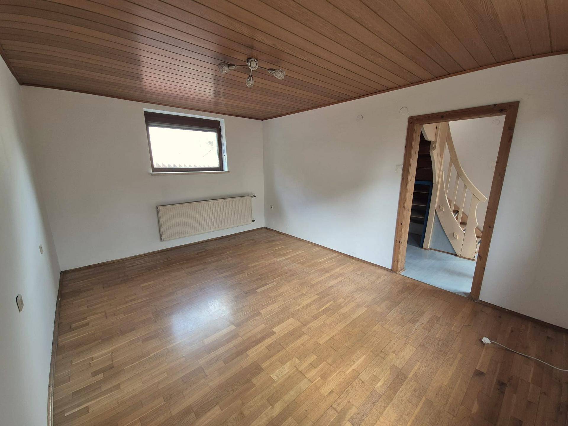 Geräumiges Zimmer mit Holzparkett, Heizkörper unter dem Fenster und Zugang zu einer Holztreppe.