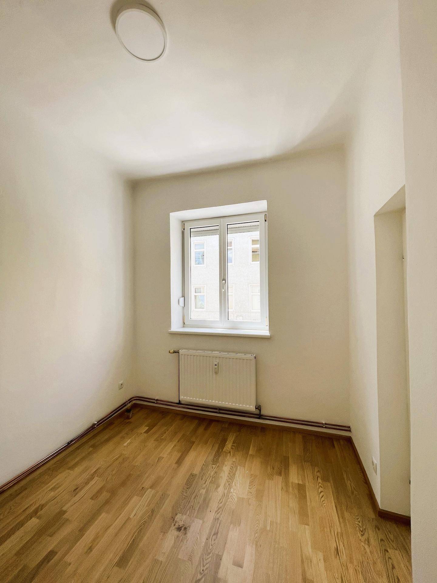 Helles Zimmer mit Parkettboden, weißen Wänden und einem Fenster mit Blick auf die Umgebung.