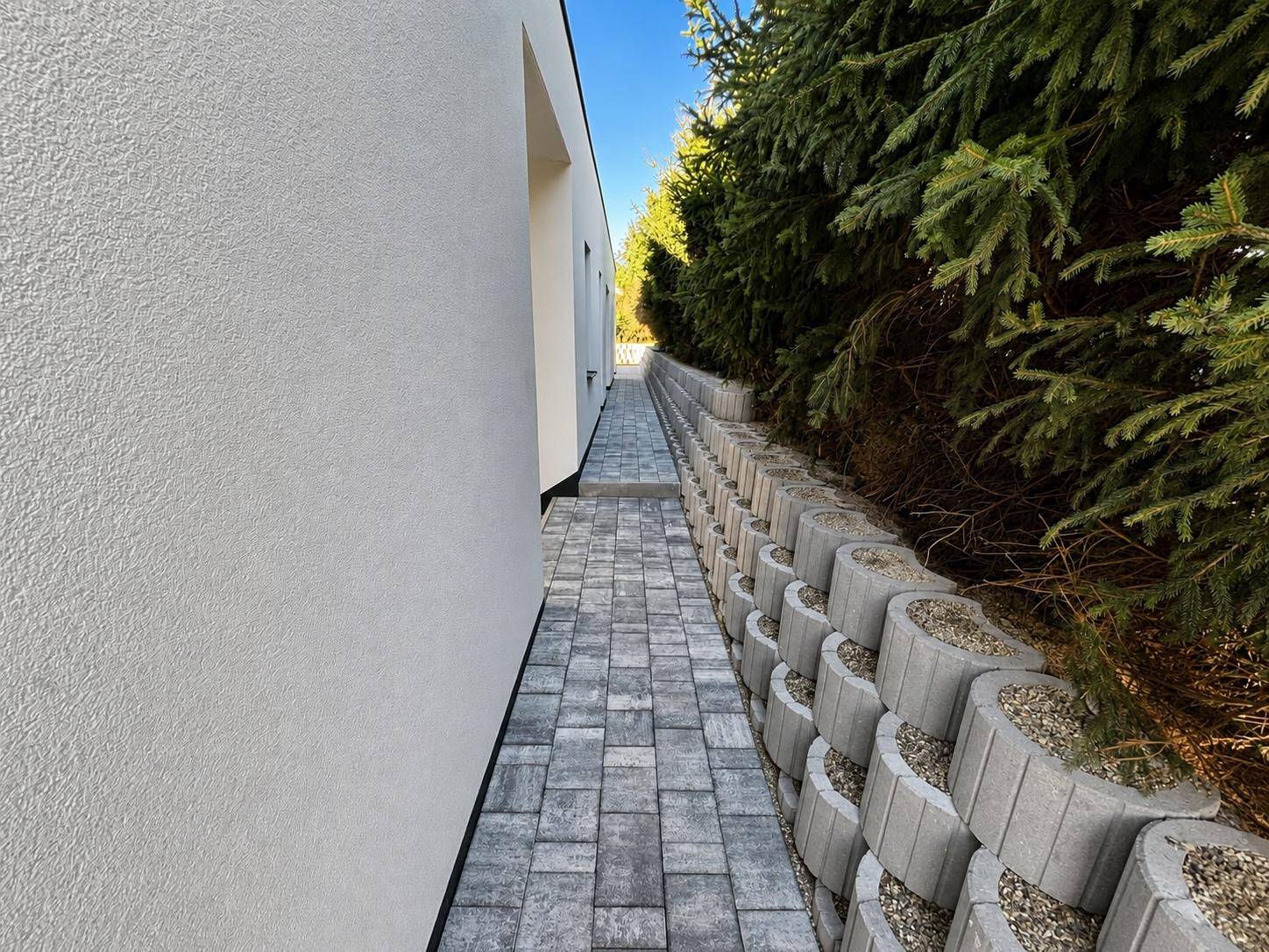 Schmaler gepflasterter Weg entlang der Hauswand mit einer begrünten Stützmauer.