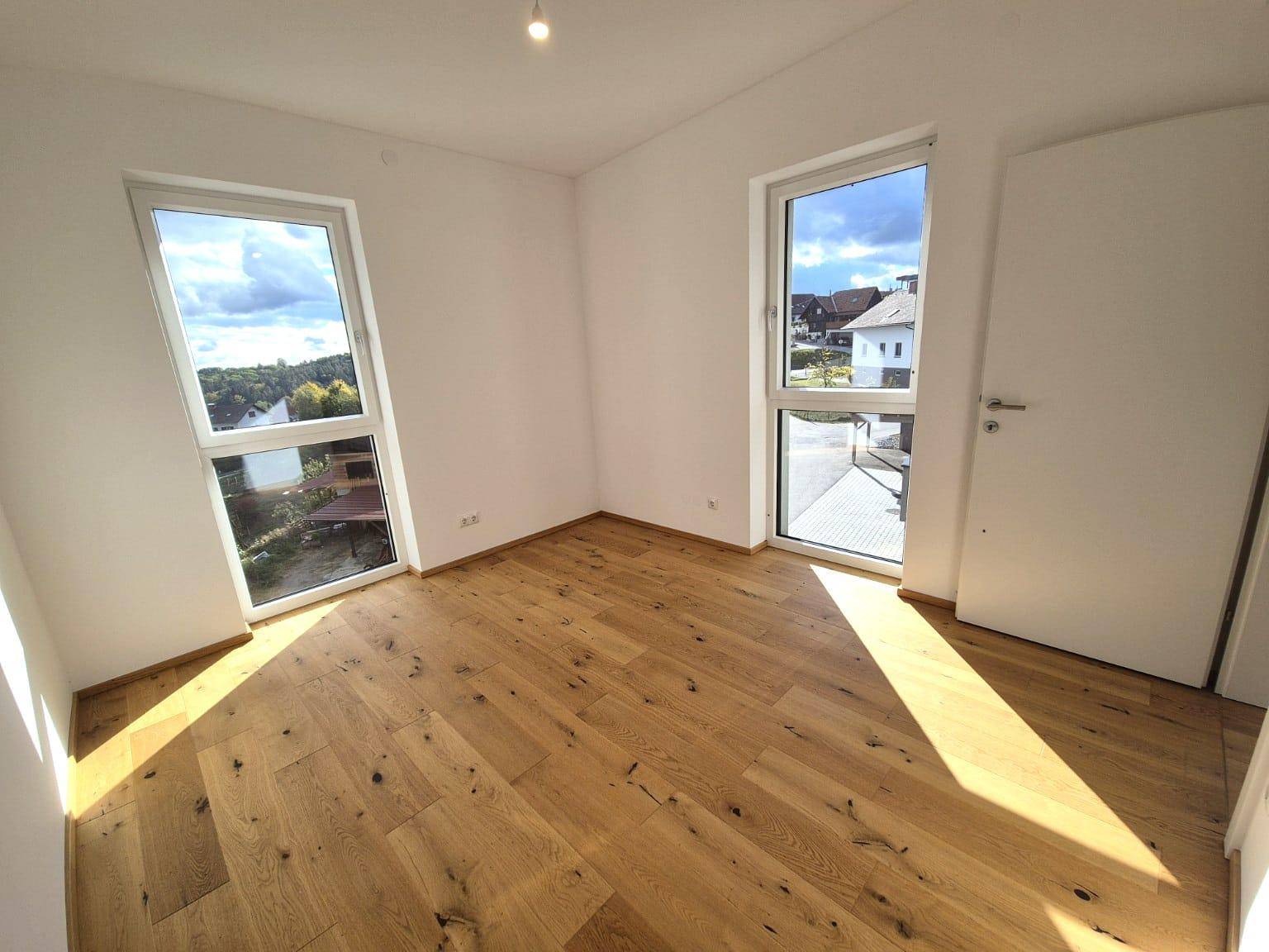 Helles Schlafzimmer mit warmem Holzdielenboden und zwei großen Fenstern, die einen Blick auf die Umgebung bieten.