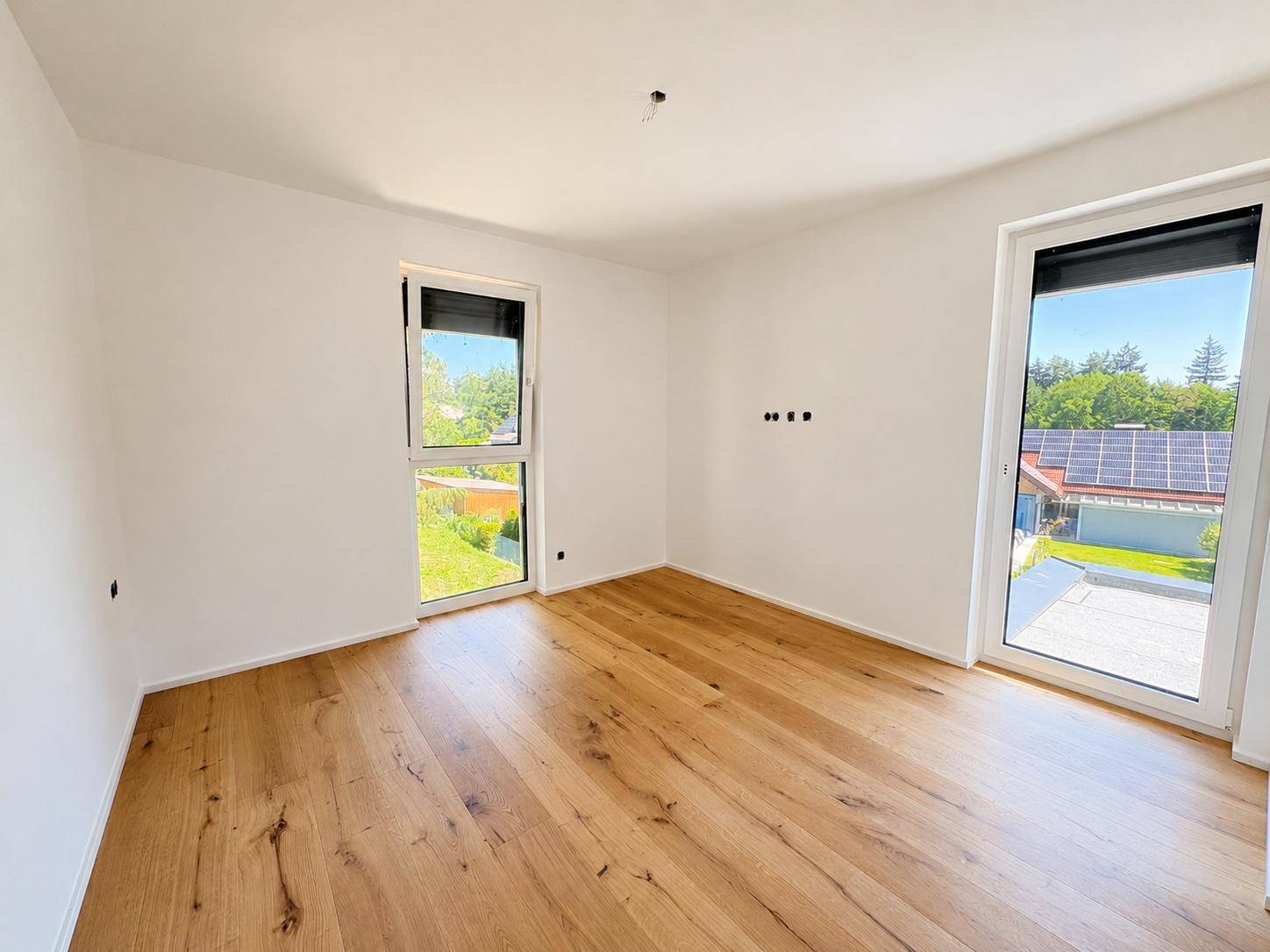 Helles Zimmer mit Holzboden, weißen Wänden und großen Fenstern, mit Blick auf eine Solaranlage.