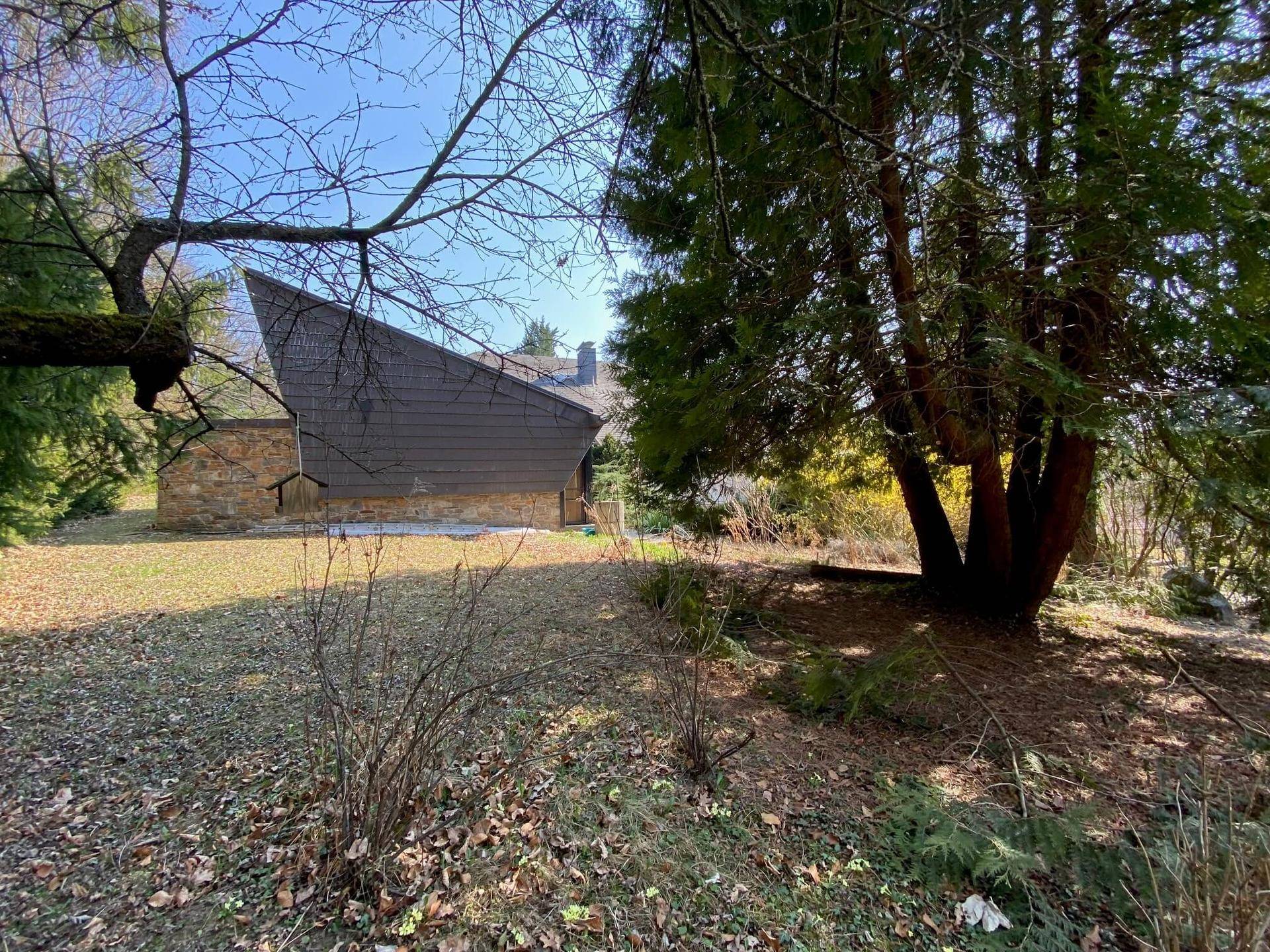 Seitenansicht des Hauses mit markanter Dachform, umgeben von Bäumen und Rasenflächen.
