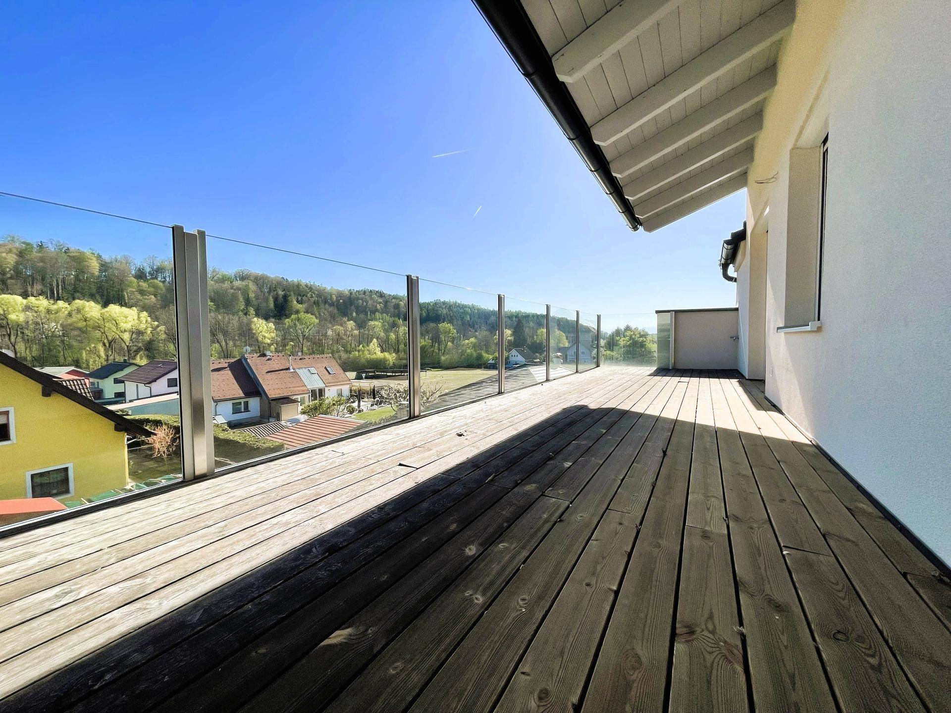 Geräumige Holzterrasse mit Glasgeländer und weitem Blick auf die grüne Umgebung.