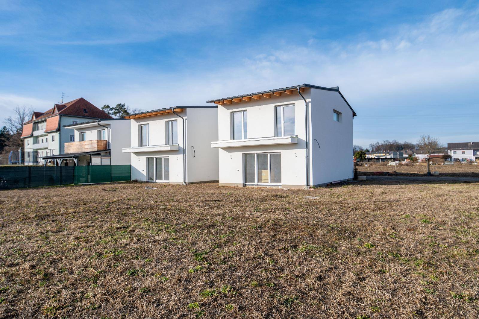 Zwei neu errichtete Häuser mit schlichter weißer Fassade unter blauem Himmel.