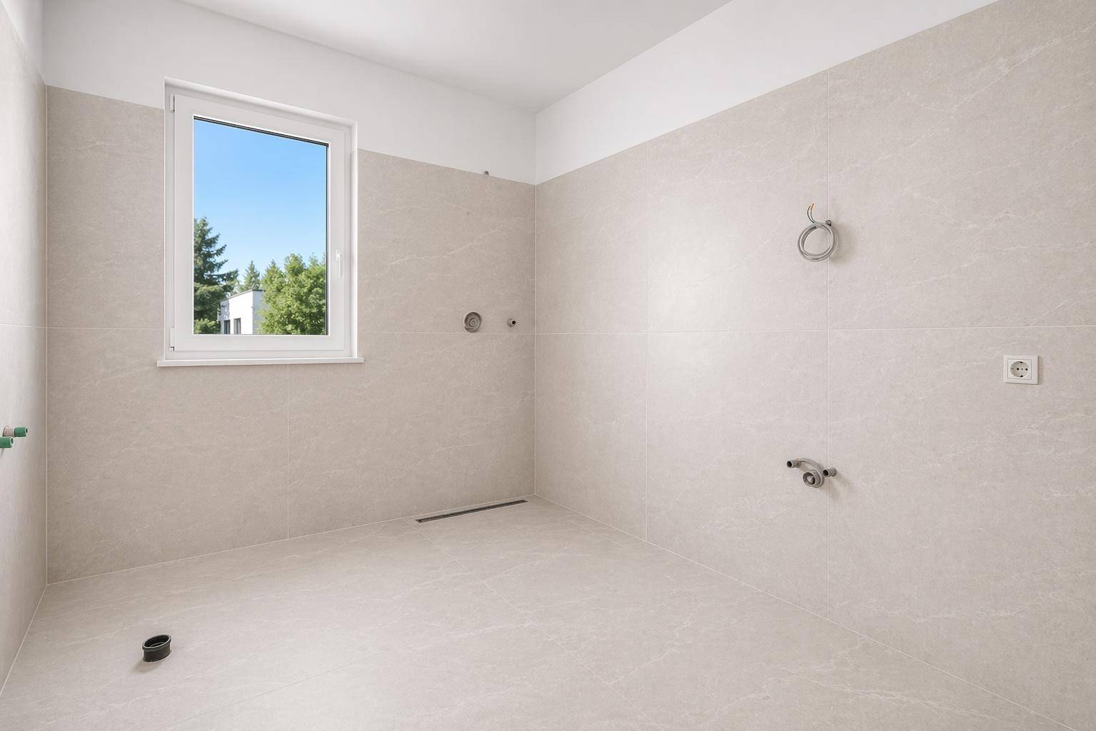 Unmöbliertes Badezimmer mit beigefarbenen Fliesen, Fenster und Anschlüssen für Sanitäranlagen.