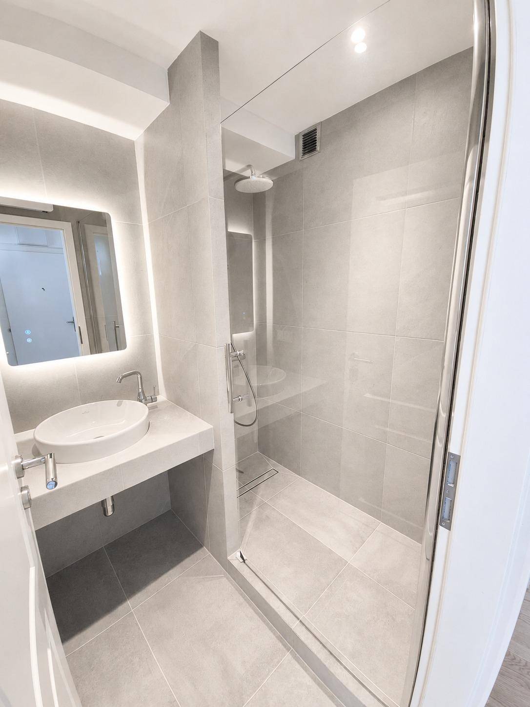 Modernes Badezimmer mit grauen Fliesen, einer begehbaren Dusche und einem beleuchteten Spiegel über dem Waschbecken.