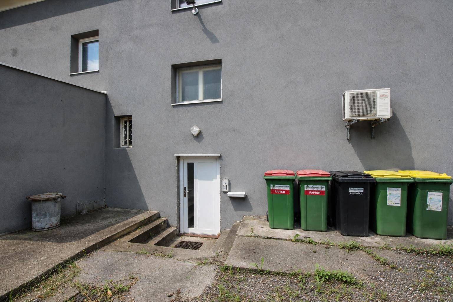 Hintereingang des Gebäudes mit einer weißen Tür, Stufen und mehreren Mülltonnen an der grauen Fassade.
