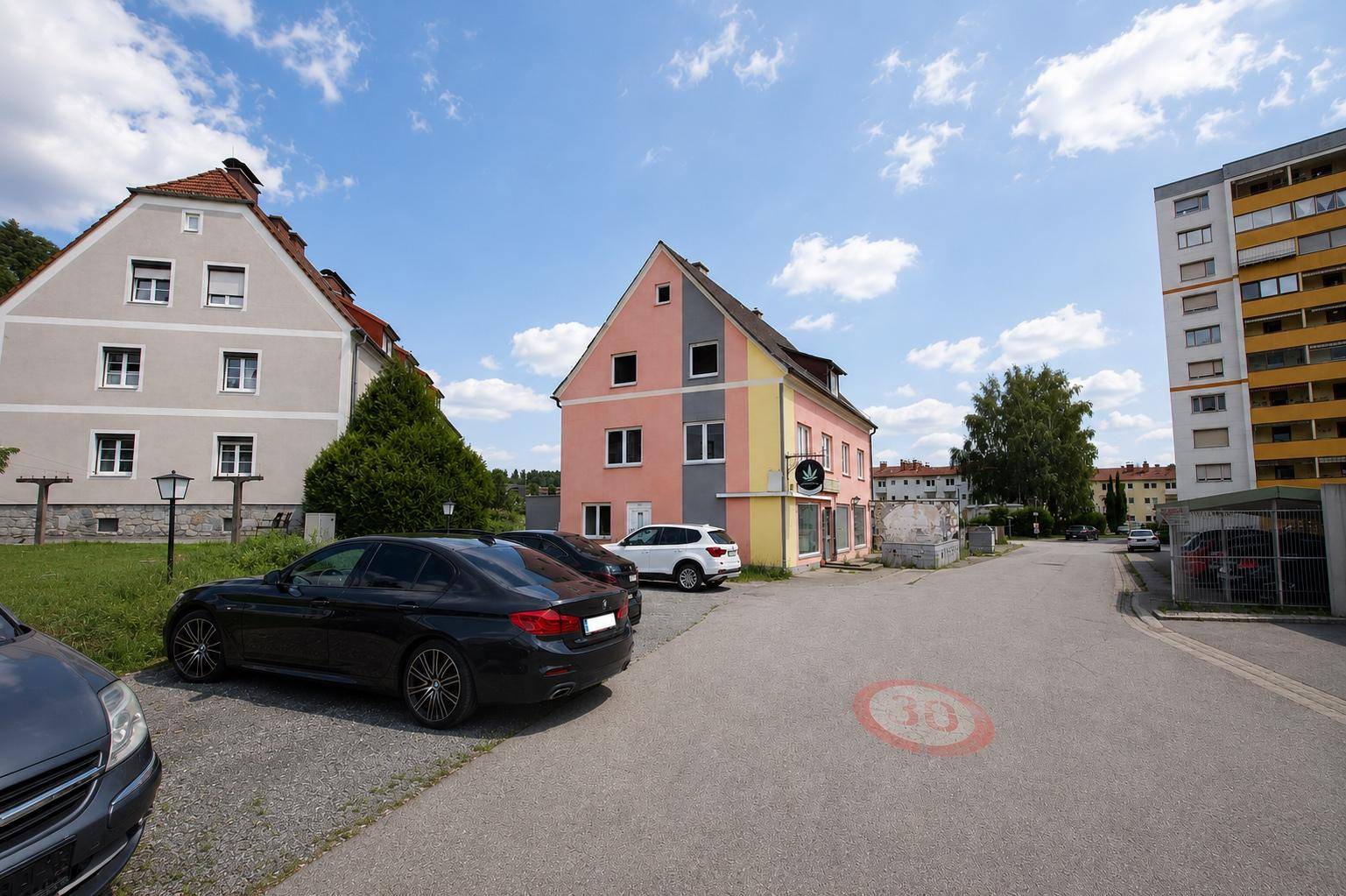 Ansicht des Mehrfamilienhauses mit rosa Fassade und umliegenden Gebäuden sowie geparkten Autos auf der Straße.