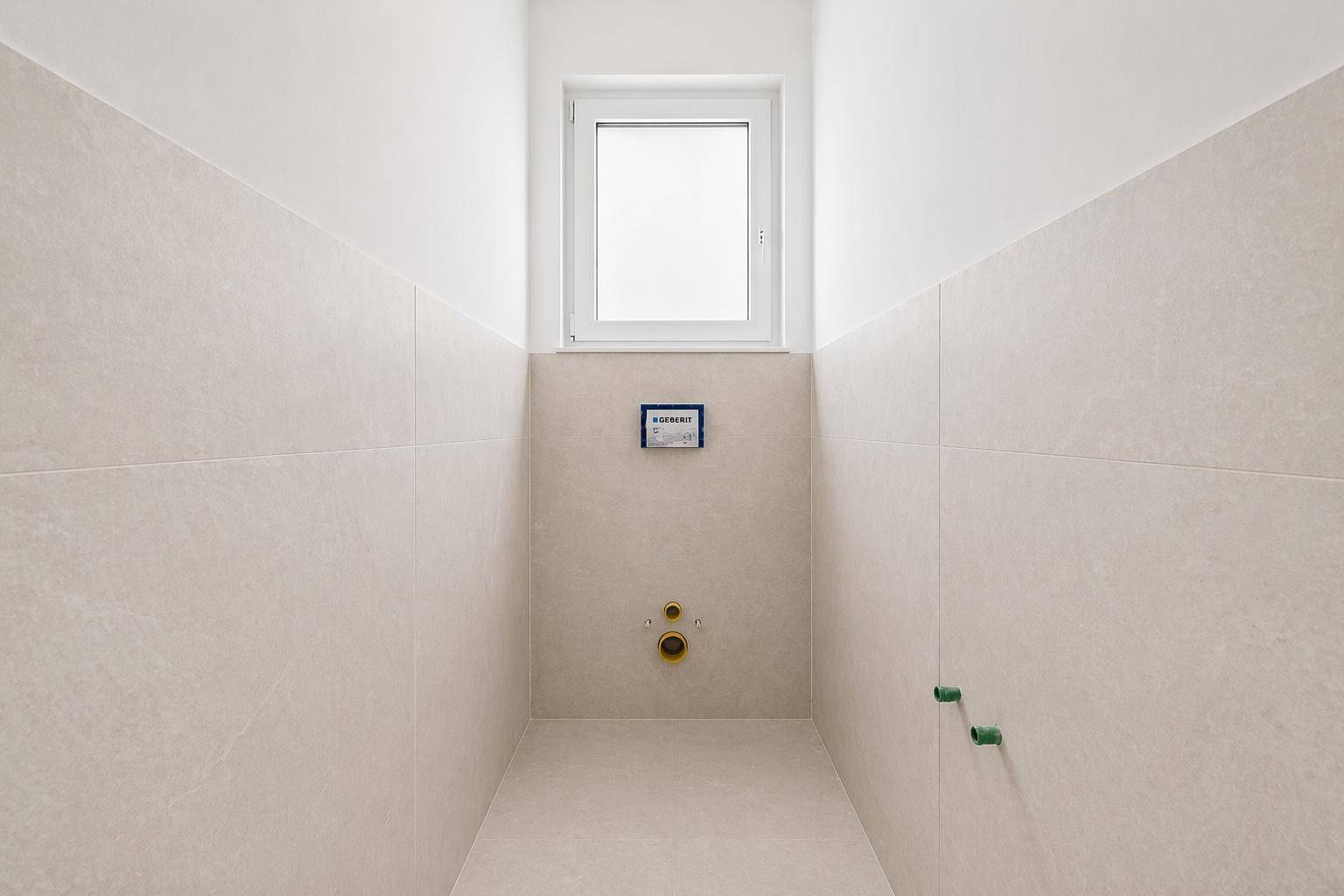 Kleines WC mit beigefarbenen Wandfliesen, Fenster und Vorbereitung für die Toiletteninstallation.