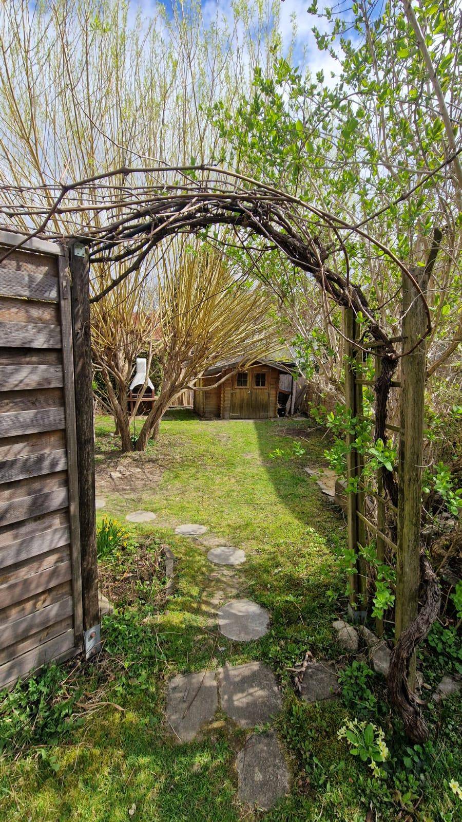 Idyllischer Gartenweg führt durch einen bewachsenen Bogen zu einem kleinen Holzschuppen.