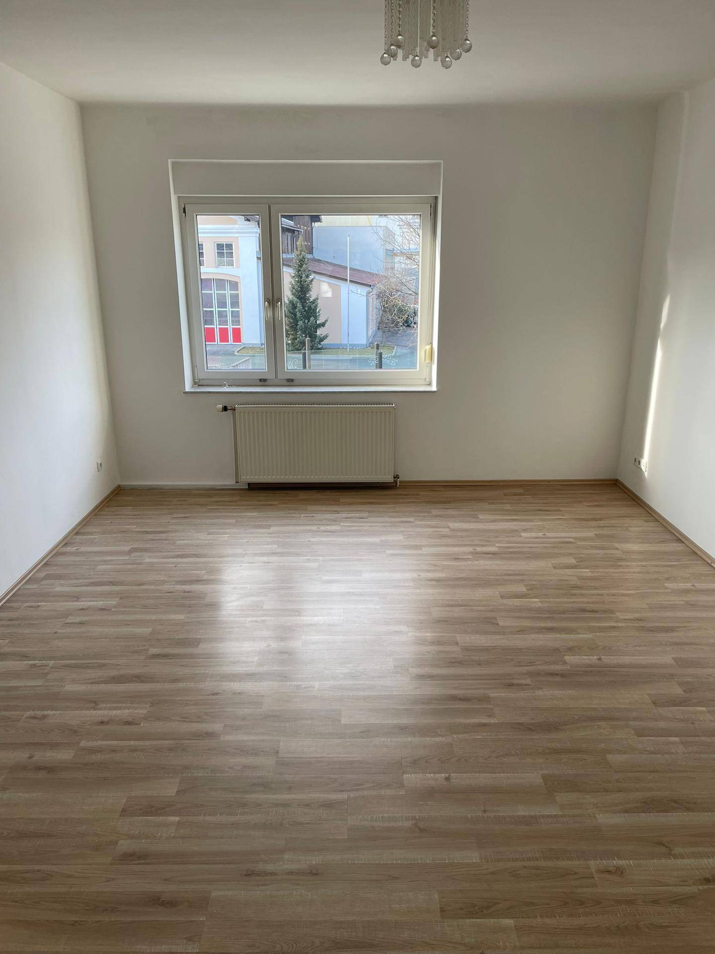 Leeres Zimmer mit hellem Laminatboden, weißen Wänden und einem Fenster mit Heizkörper darunter.