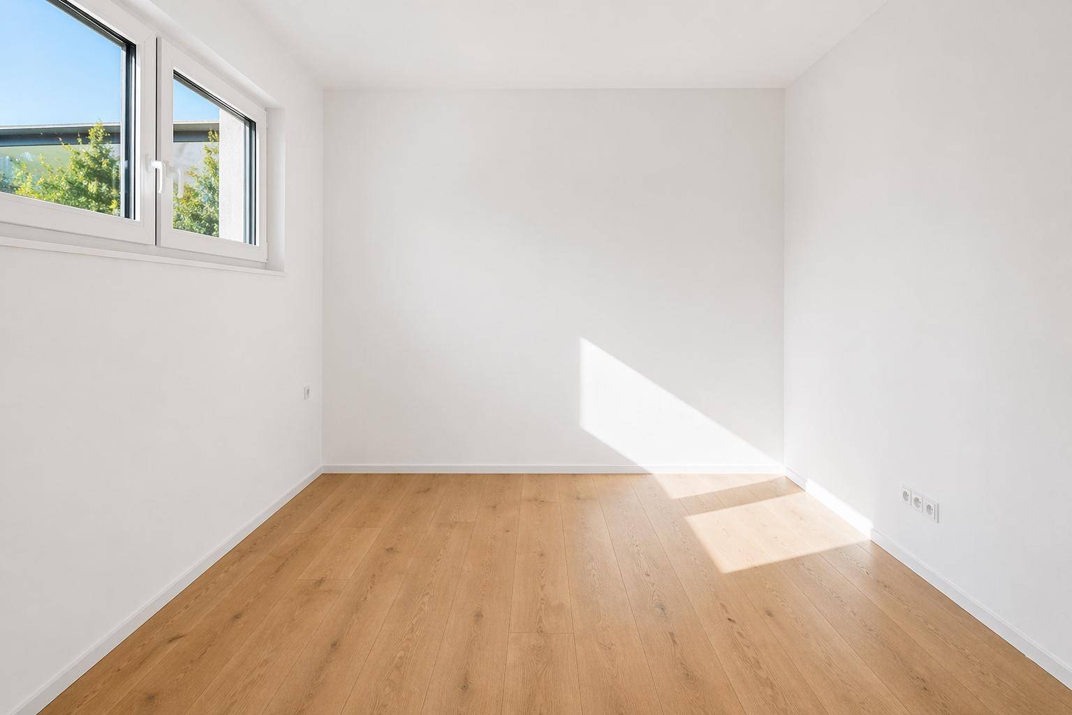 Leeres Zimmer mit hellem Holzboden und einem Fenster, das viel Sonnenlicht hereinlässt.