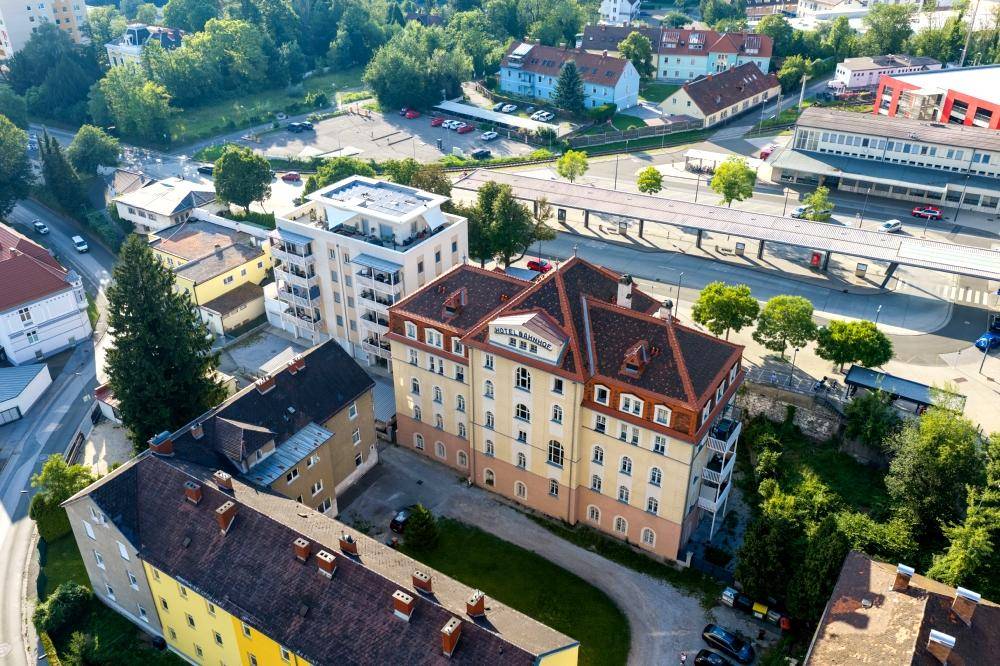 Luftaufnahme eines historischen Hotels und eines modernen Wohngebäudes in einer grünen Stadtlandschaft.
