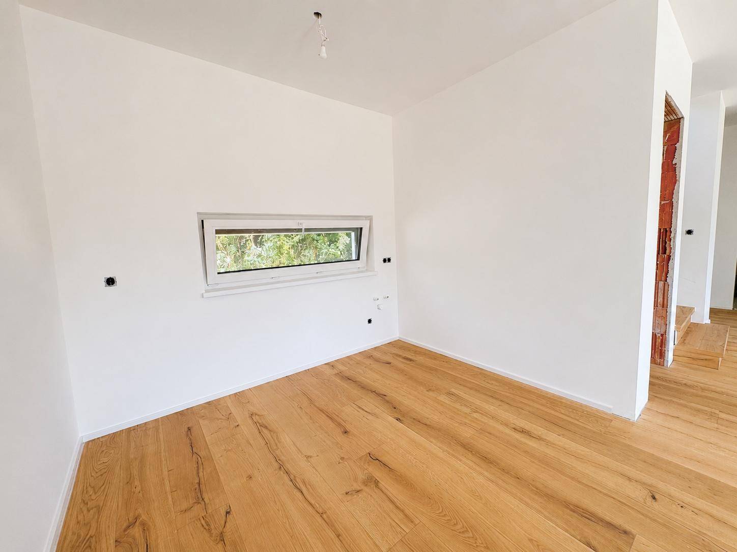 Geräumiges Zimmer mit Holzboden und einem breiten Fenster, das den Raum hell erscheinen lässt.