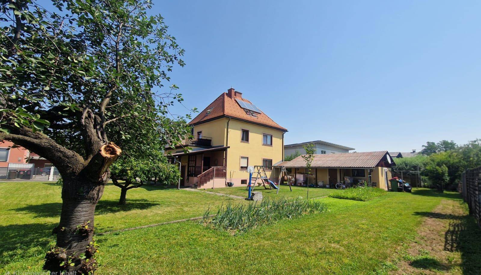 Großer Garten mit viel Grünfläche, Bäumen und einem kleinen Spielplatz vor dem gelben Haus.