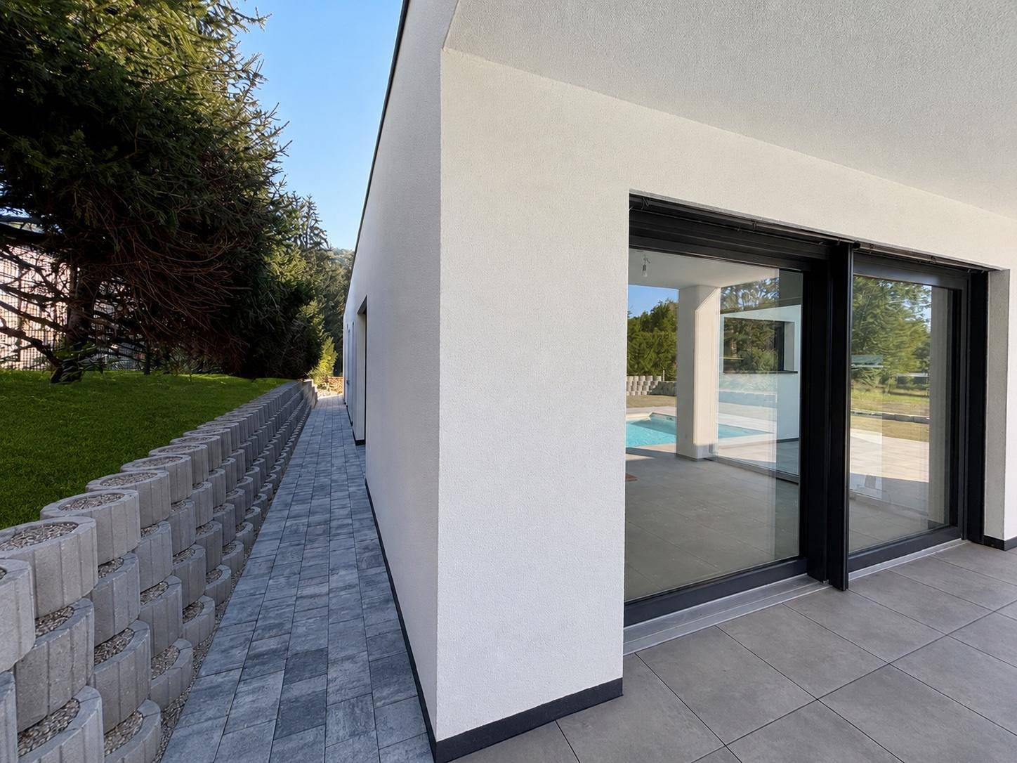 Seitenansicht des Bungalows mit gepflastertem Weg, Stützmauer und großen Glasschiebetüren.