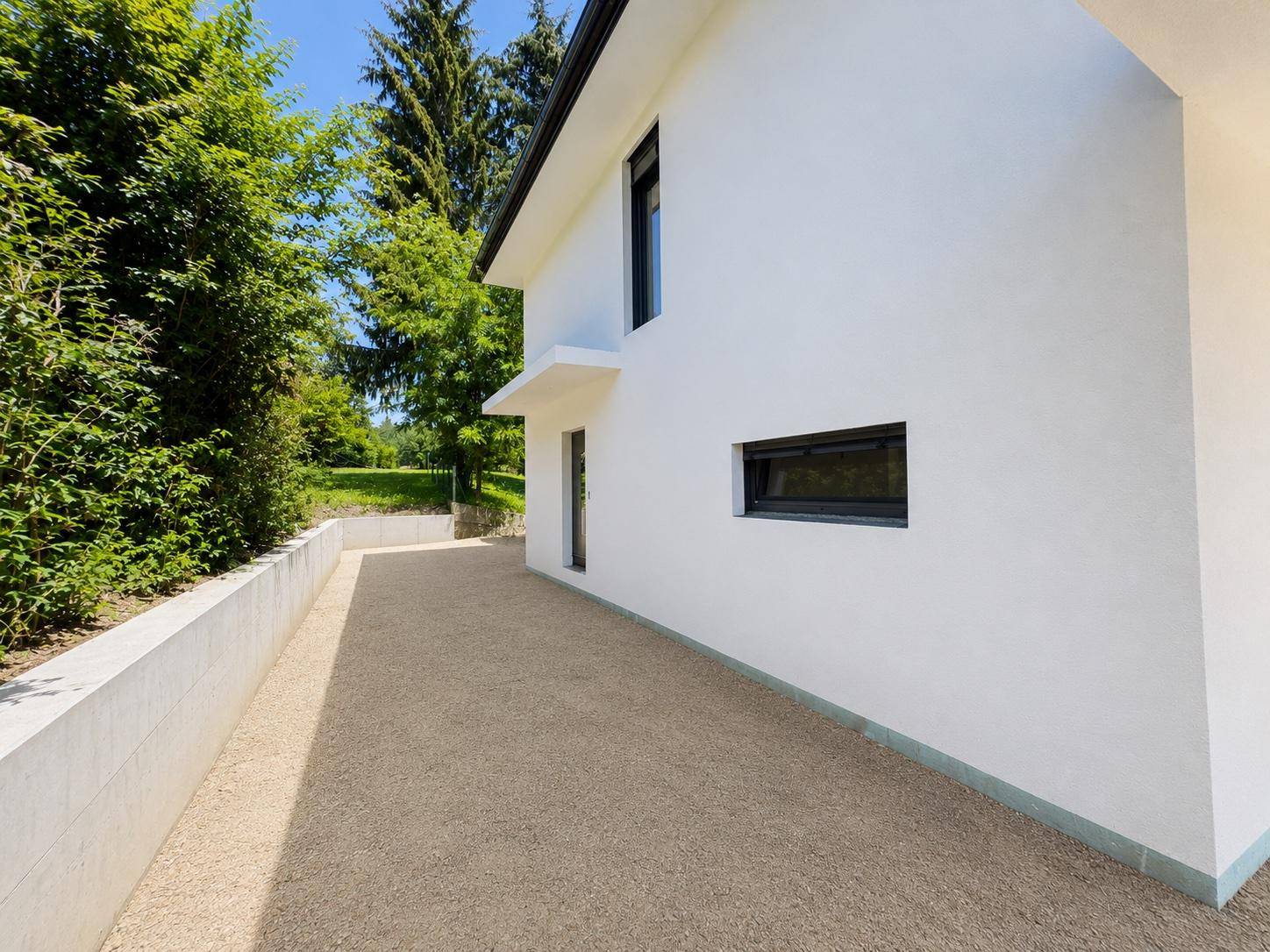 Seitlicher Weg entlang des Hauses mit Kiesfläche, Stützmauer und grüner Bepflanzung.