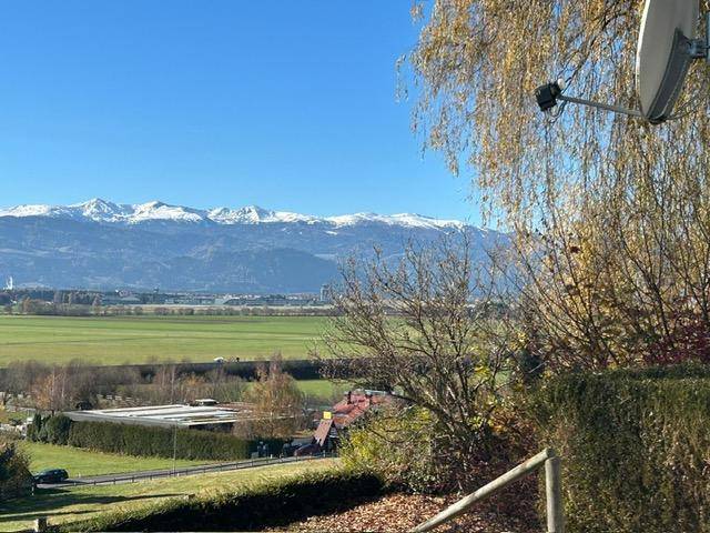 Weitläufiger Blick über grüne Felder und schneebedeckte Berge unter strahlend blauem Himmel.