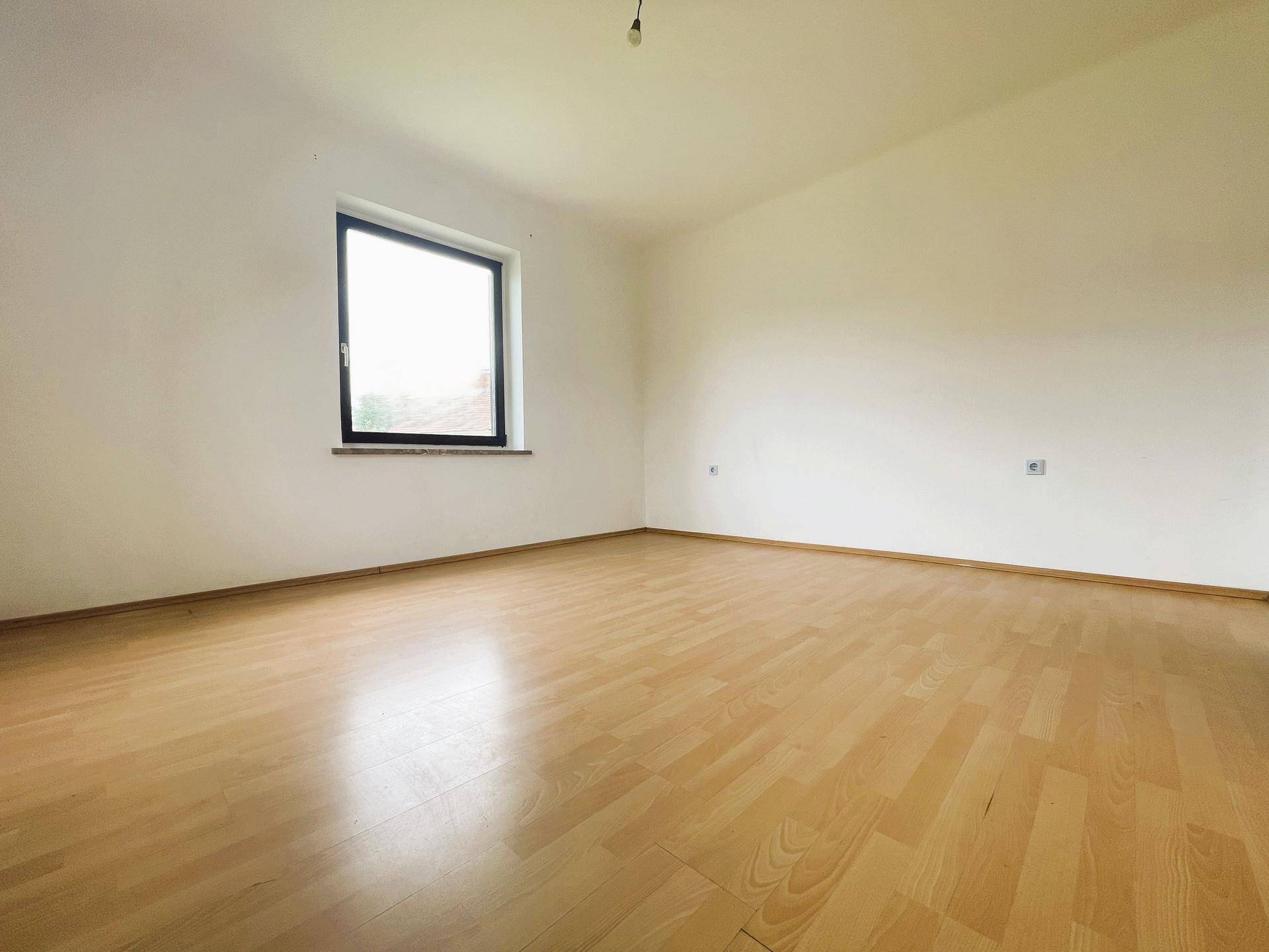 Lichtdurchfluteter Raum mit einem Fenster, hellem Laminatboden und weißen Wänden.