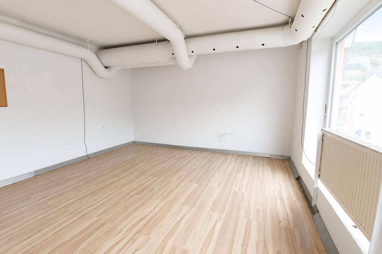 Leerer Raum mit hellem Laminatboden, weißen Wänden und sichtbaren Lüftungsrohren an der Decke.