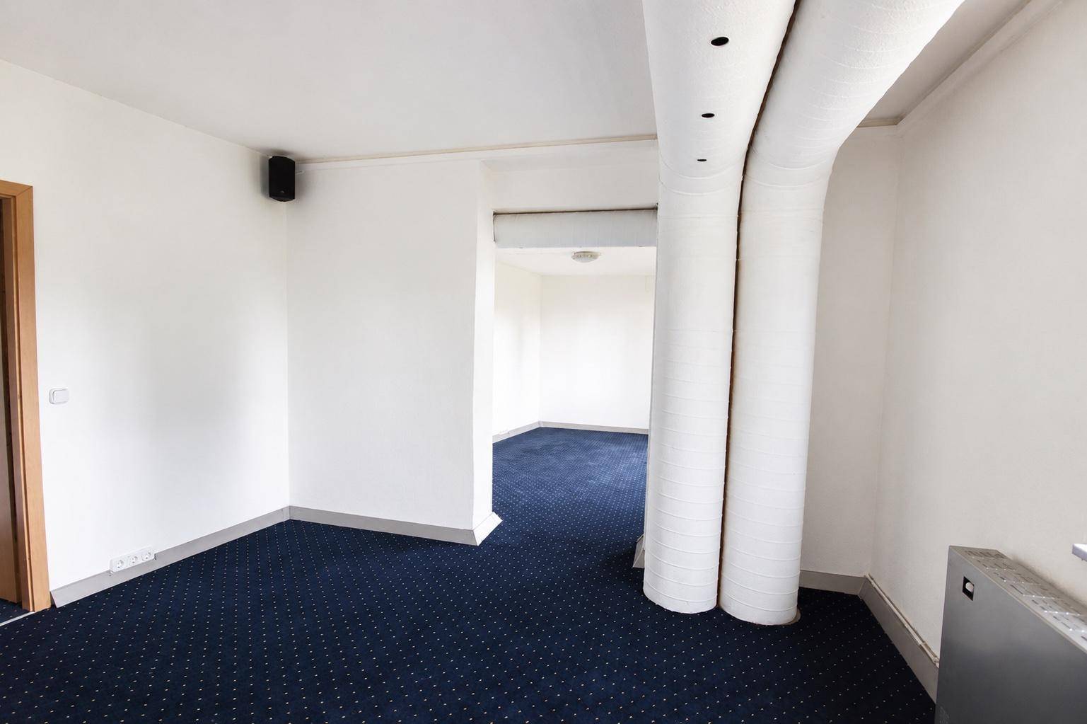 Geräumiger Raum mit blauem Teppichboden, weißen Wänden und sichtbaren Lüftungsrohren an der Decke.