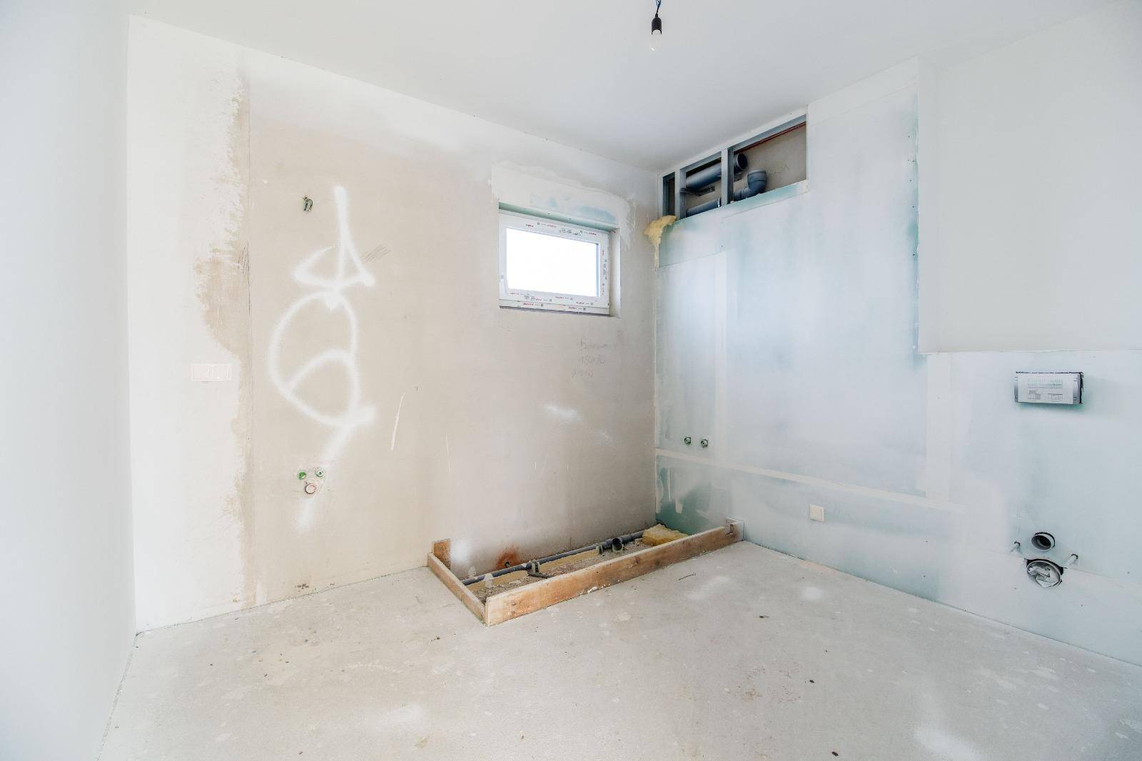 Unfertiges Badezimmer im Rohbau mit Fenster und vorbereiteten Anschlüssen für Sanitäranlagen.