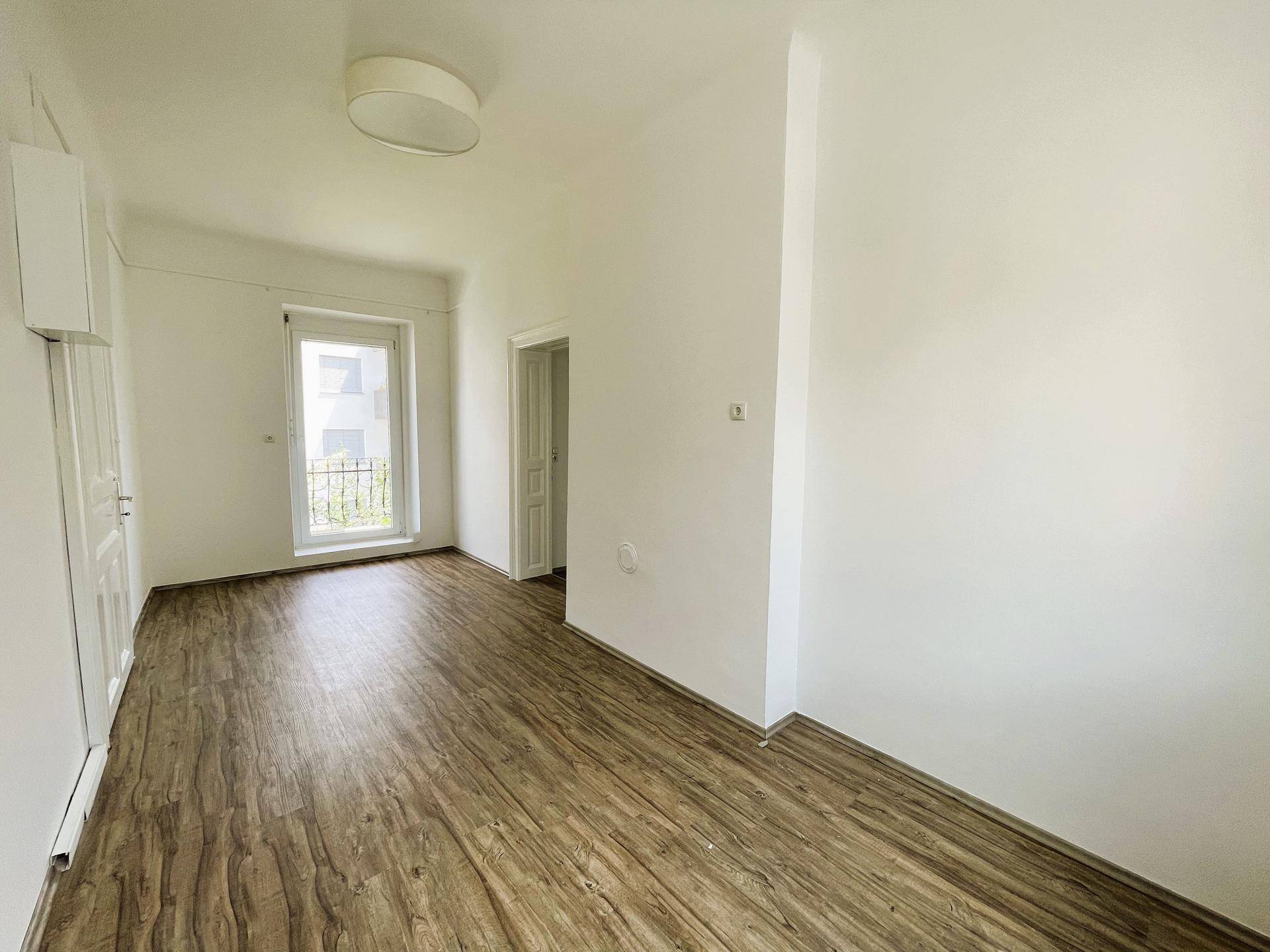 Leeres Zimmer mit Laminatboden und Zugang zu einem kleinen Balkon, bietet viel Tageslicht.