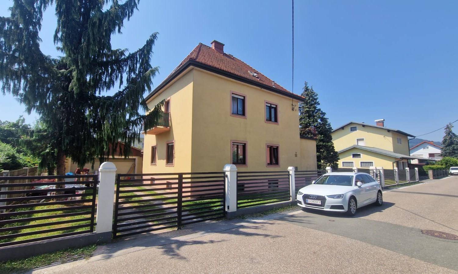 Ansicht des gelben Hauses von der Straße mit einem modernen Zaun und geparktem Auto.