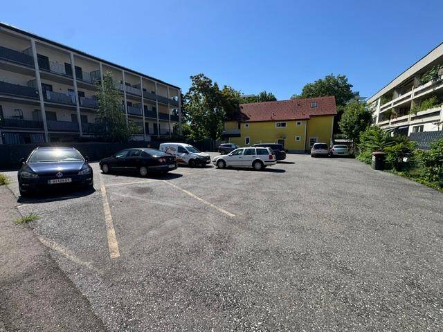 Weitläufiger Parkplatz vor einem gelben Gebäude und modernen Wohnkomplexen an einem sonnigen Tag.