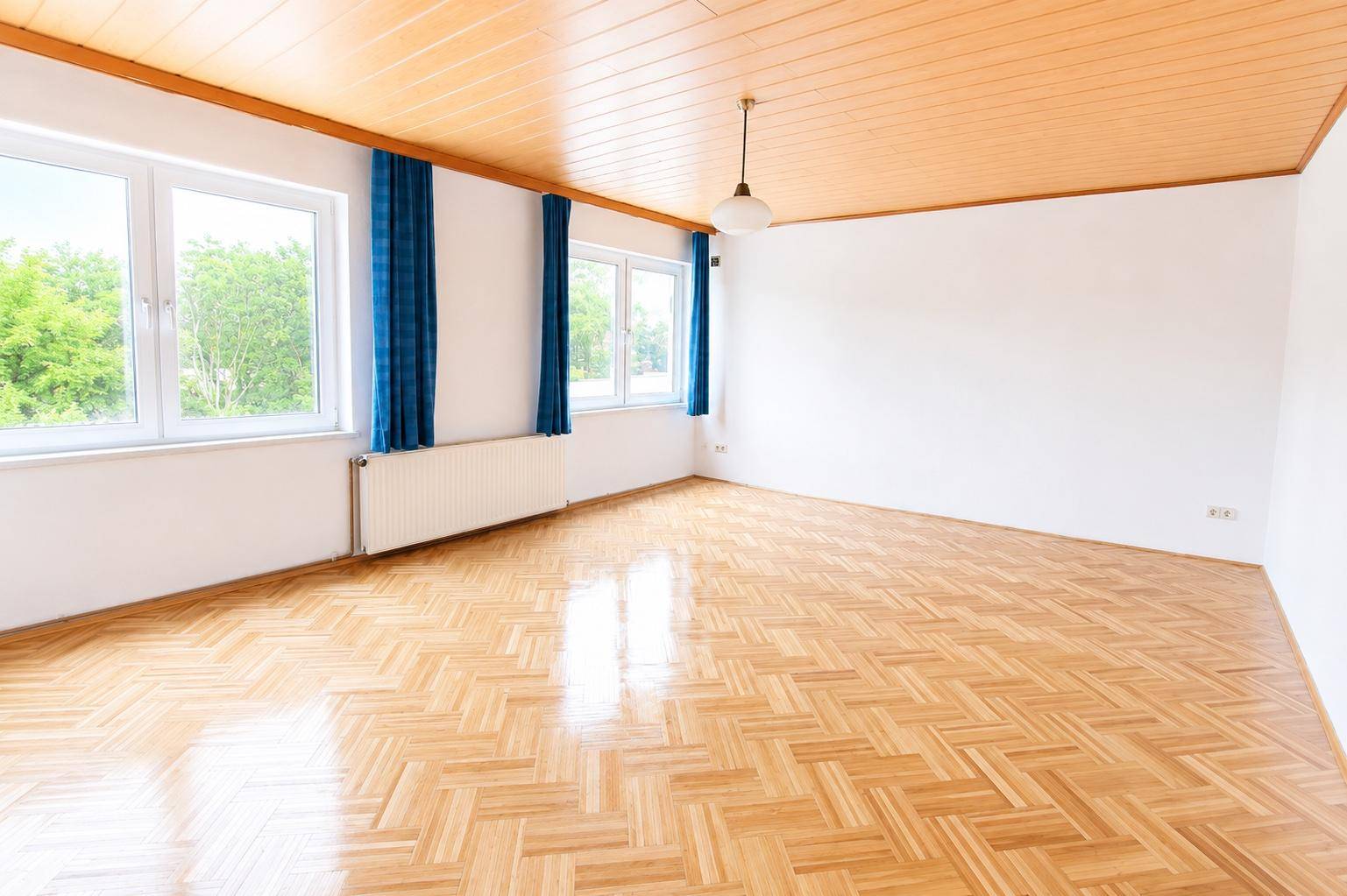 Geräumiger Raum mit hellem Holzparkettboden, weißen Wänden und blauen Vorhängen an den Fenstern.