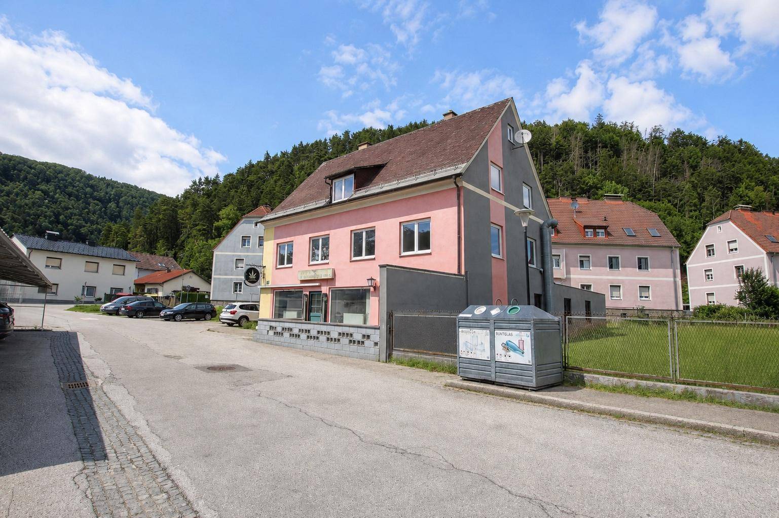 Mehrfamilienhaus mit rosa und grauer Fassade an einer Straße, umgeben von grünen Hügeln und Bäumen.