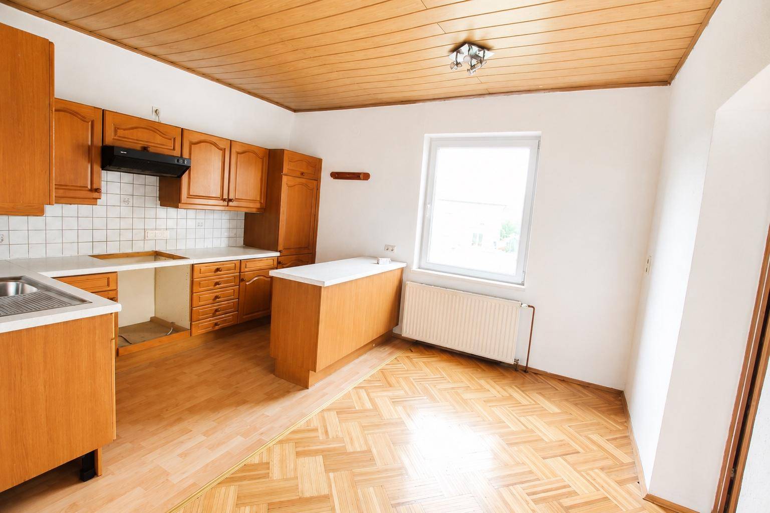 Küche mit Holzschränken, hellem Parkettboden und einem Fenster, das viel Licht hereinlässt.
