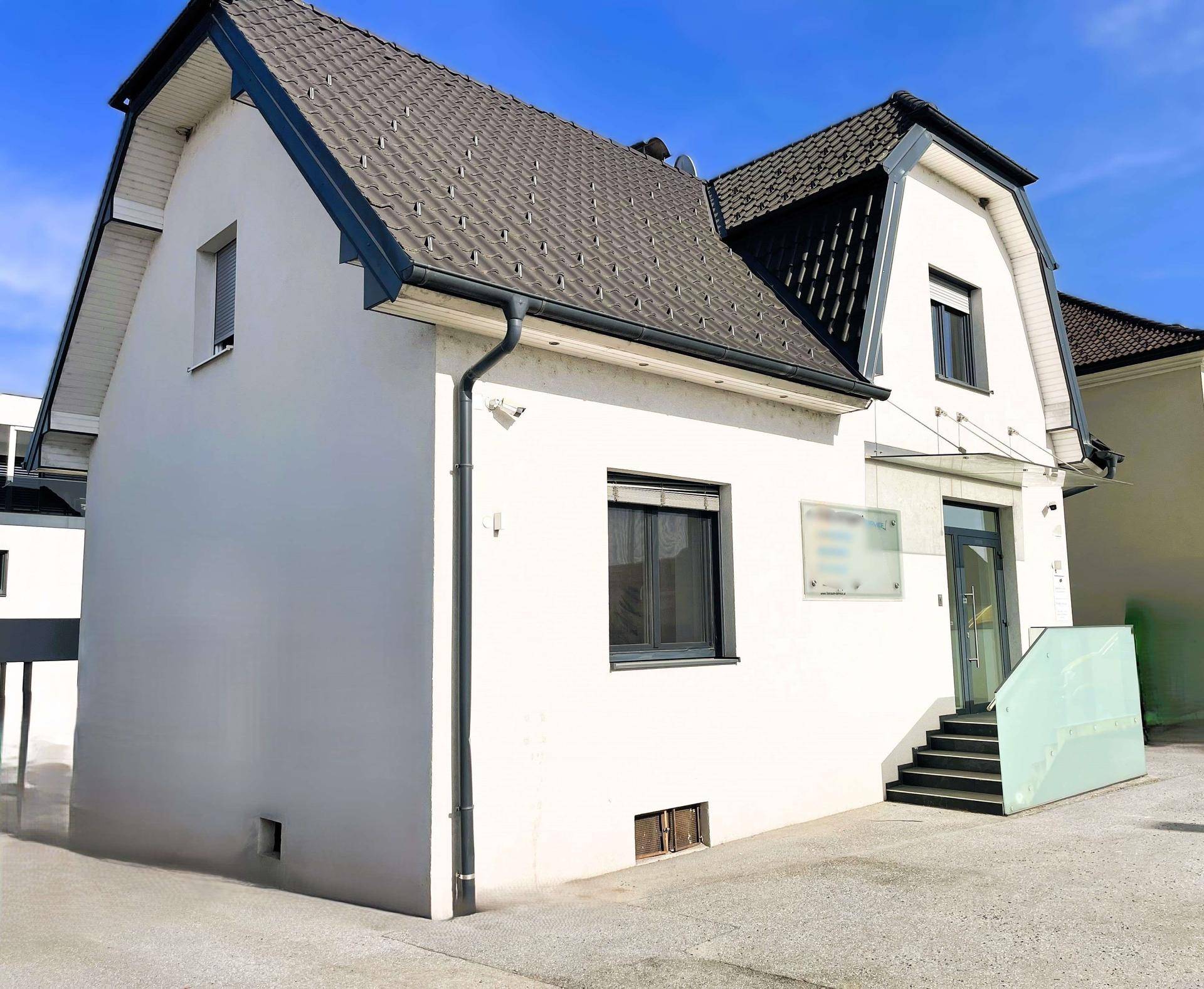 Seitenansicht eines weißen Gebäudes mit dunklem Satteldach und gepflastertem Vorplatz.