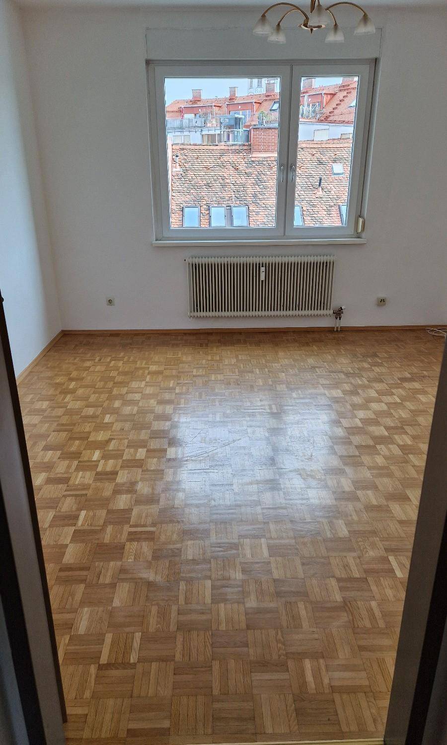 Helles Zimmer mit Parkettboden und einem Fenster, das einen Blick über die Dächer bietet.