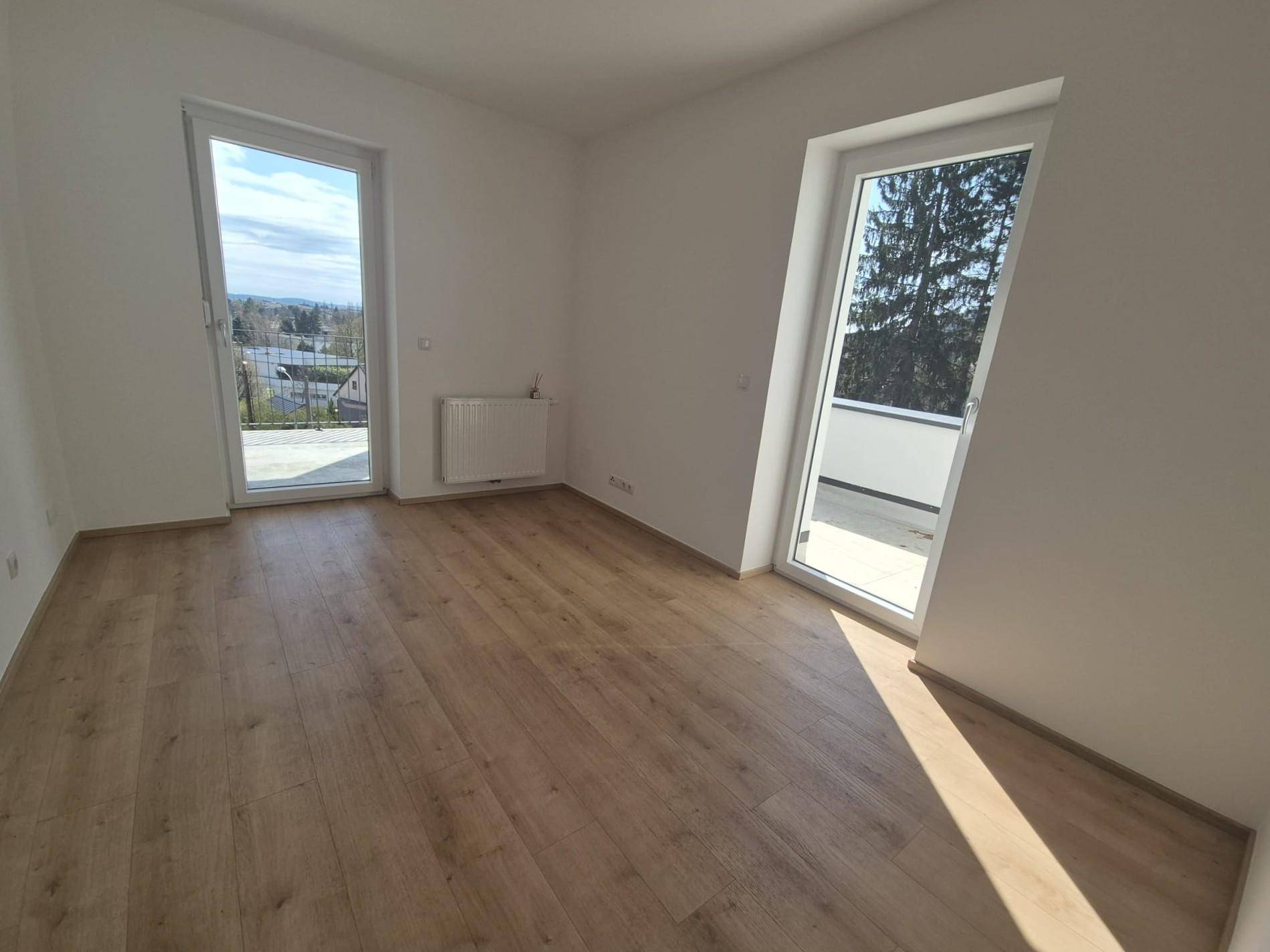 Heller Wohnraum mit Laminatboden und großen Fenstern, die Zugang zu einem Außenbereich bieten.