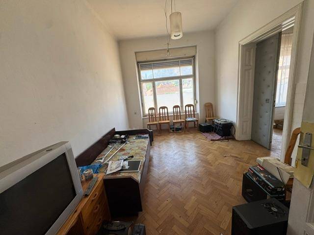 Geräumiges Wohnzimmer mit Fischgrätparkett und großem Fenster, renovierungsbedürftig.