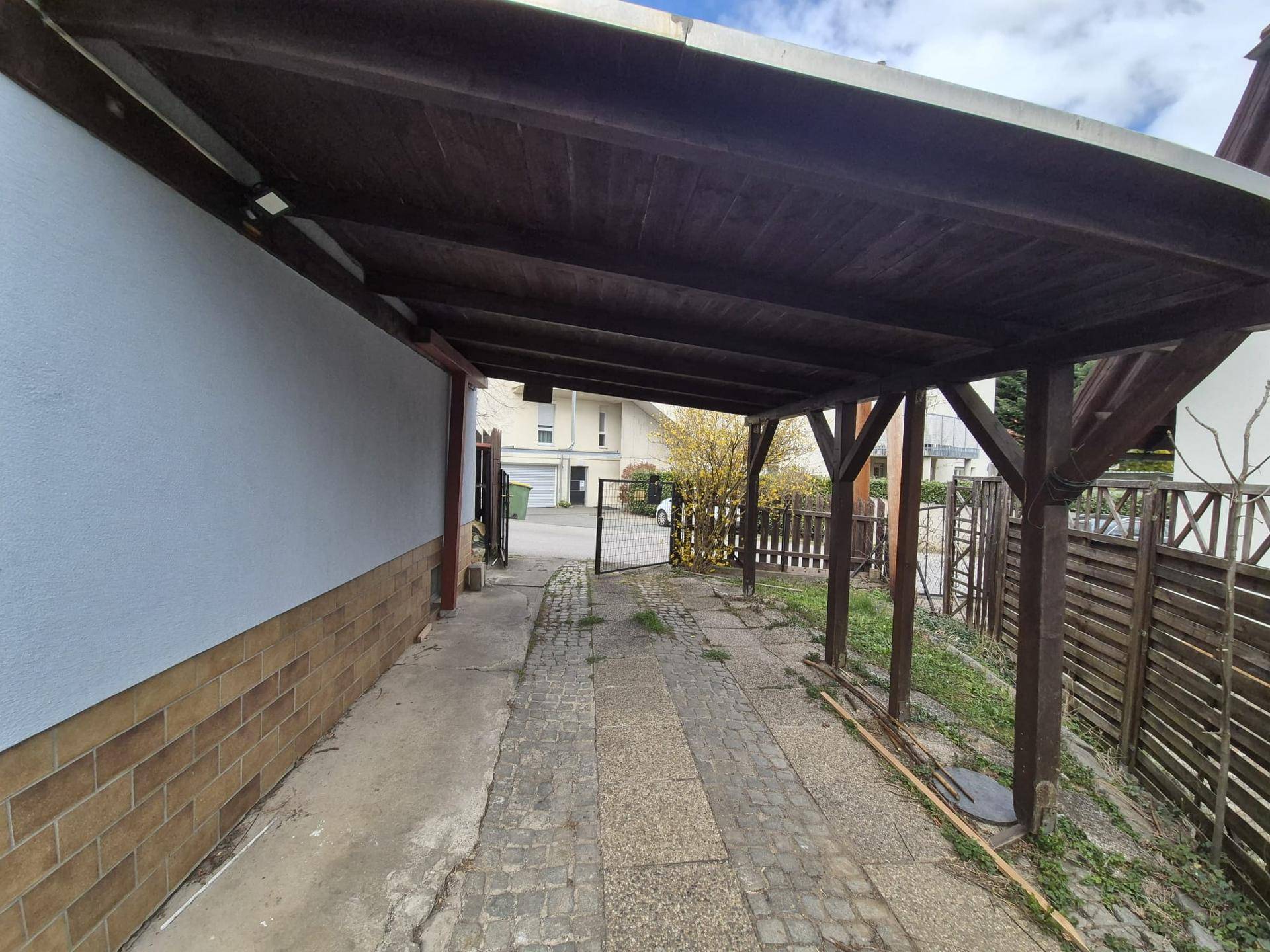Überdachter Carport mit Holzbalken und gepflasterter Zufahrt neben dem Haus.