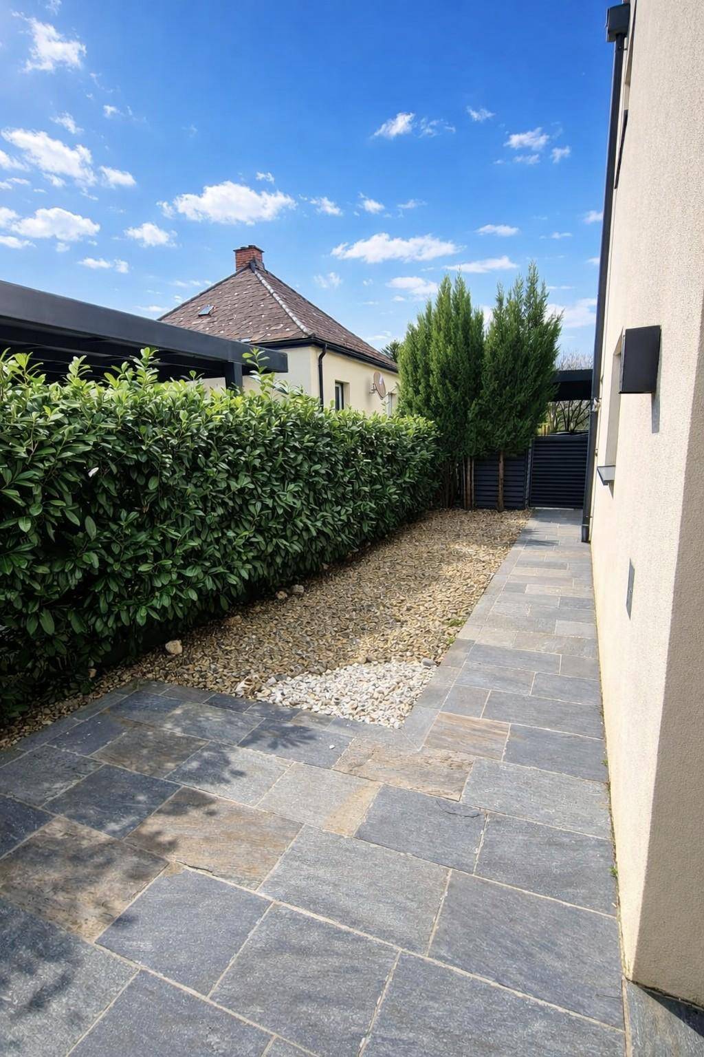 Gepflegter Gartenweg mit Steinplatten und hoher Hecke entlang des Gebäudes.