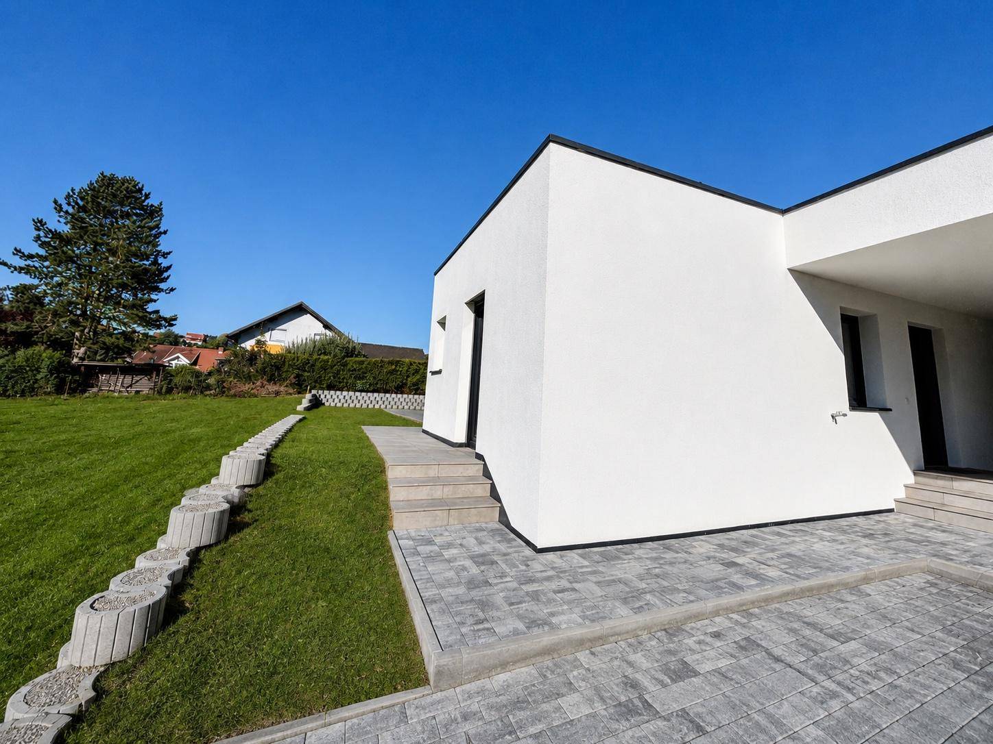 Seitenansicht des modernen Bungalows mit Eingangsbereich, gepflastertem Weg und Rasenfläche.