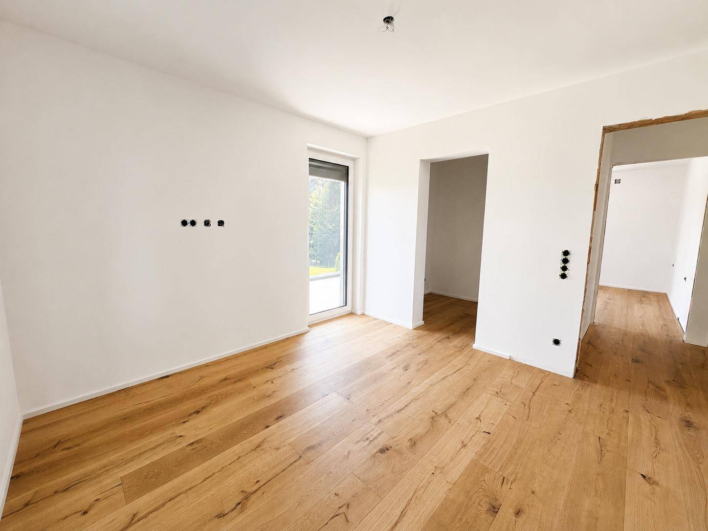 Heller Raum mit Holzboden, weißen Wänden und einem Fenster, angrenzend an weitere Räume.