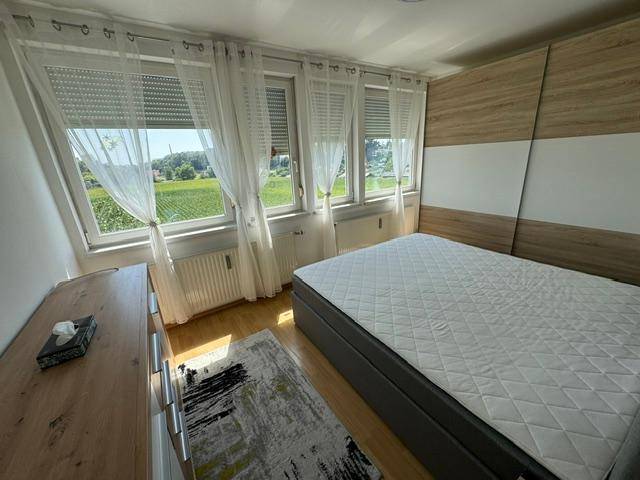 Helles Schlafzimmer mit Doppelbett, Holzschrank und weitem Blick ins Grüne durch große Fenster.