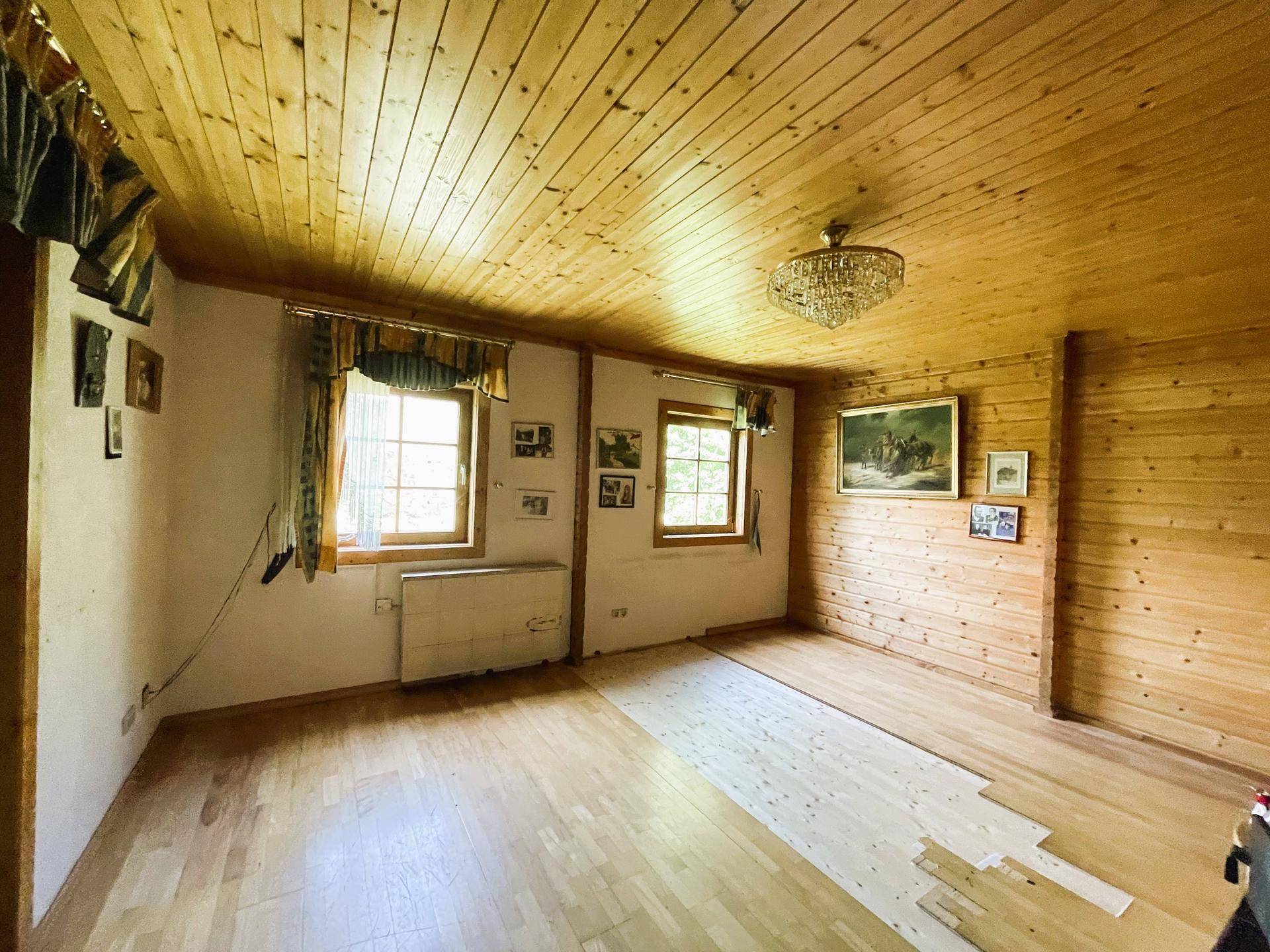 Helles Wohnzimmer mit Holzwänden, Holzdecke und zwei Fenstern, teilweise mit neuem Boden.
