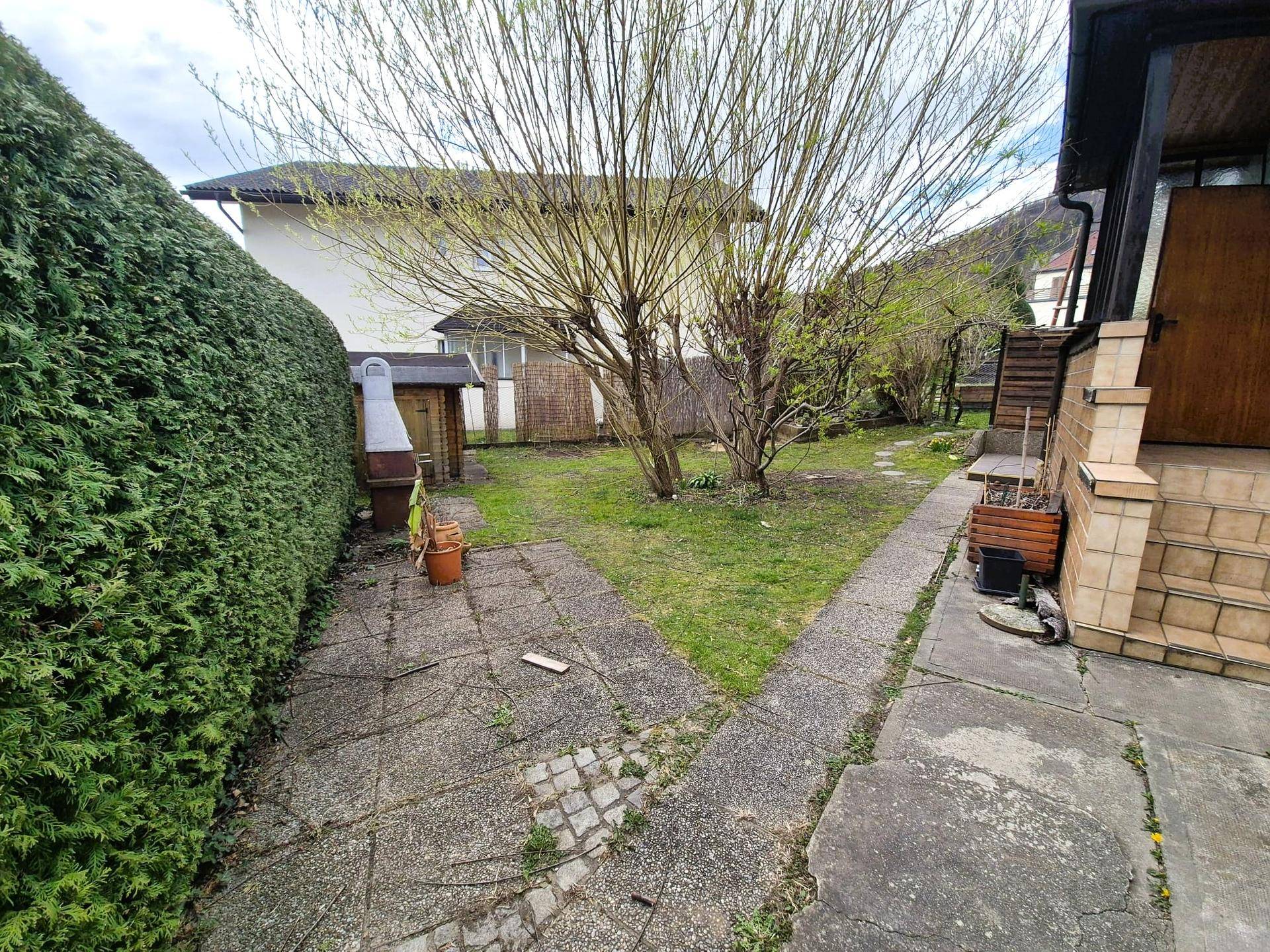 Garten mit Hecke, gepflastertem Weg und einem kleinen Grillplatz.