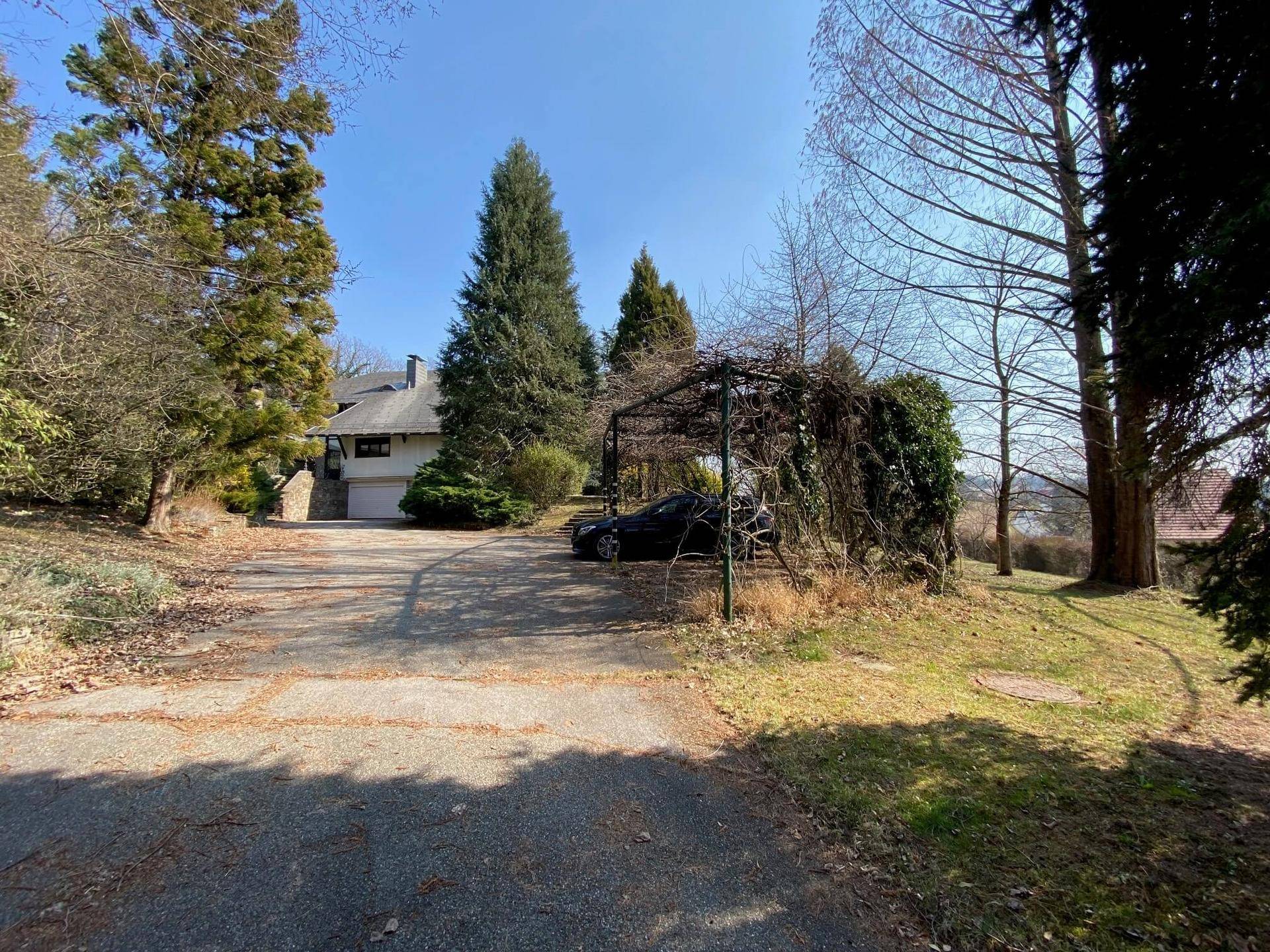 Hausansicht mit großer Einfahrt, Carport und üppiger Vegetation im sonnigen Außenbereich.