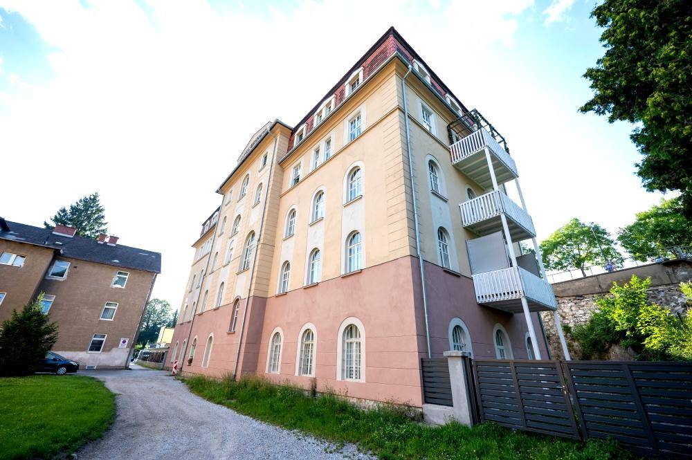 Historisches Hotelgebäude mit seitlichem Anbau, Rasenfläche und Bäumen unter blauem Himmel.