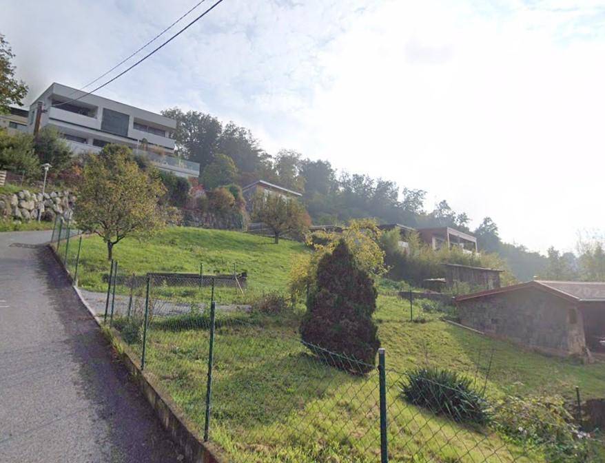 Straße entlang eines Hangs mit modernen Gebäuden und grünen Grundstücken in einer hügeligen Umgebung.