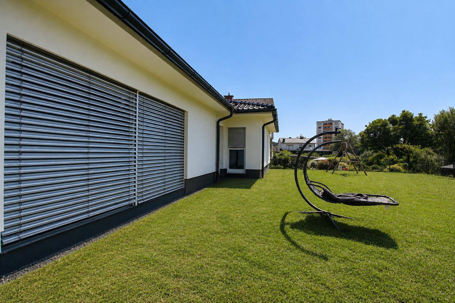 Seitliche Ansicht des Bungalows mit großen Fenstern und einer modernen Hängeliege im Garten.