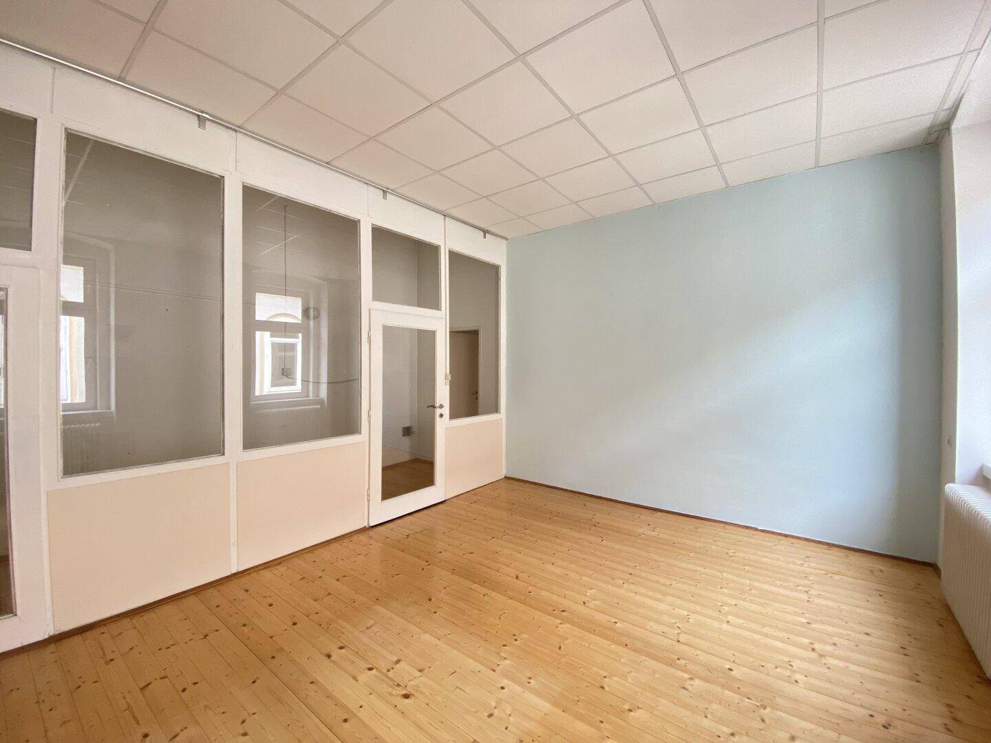 Heller Büroraum mit Holzplankenboden, Rasterdecke und einer Wand mit Glasabtrennungen.