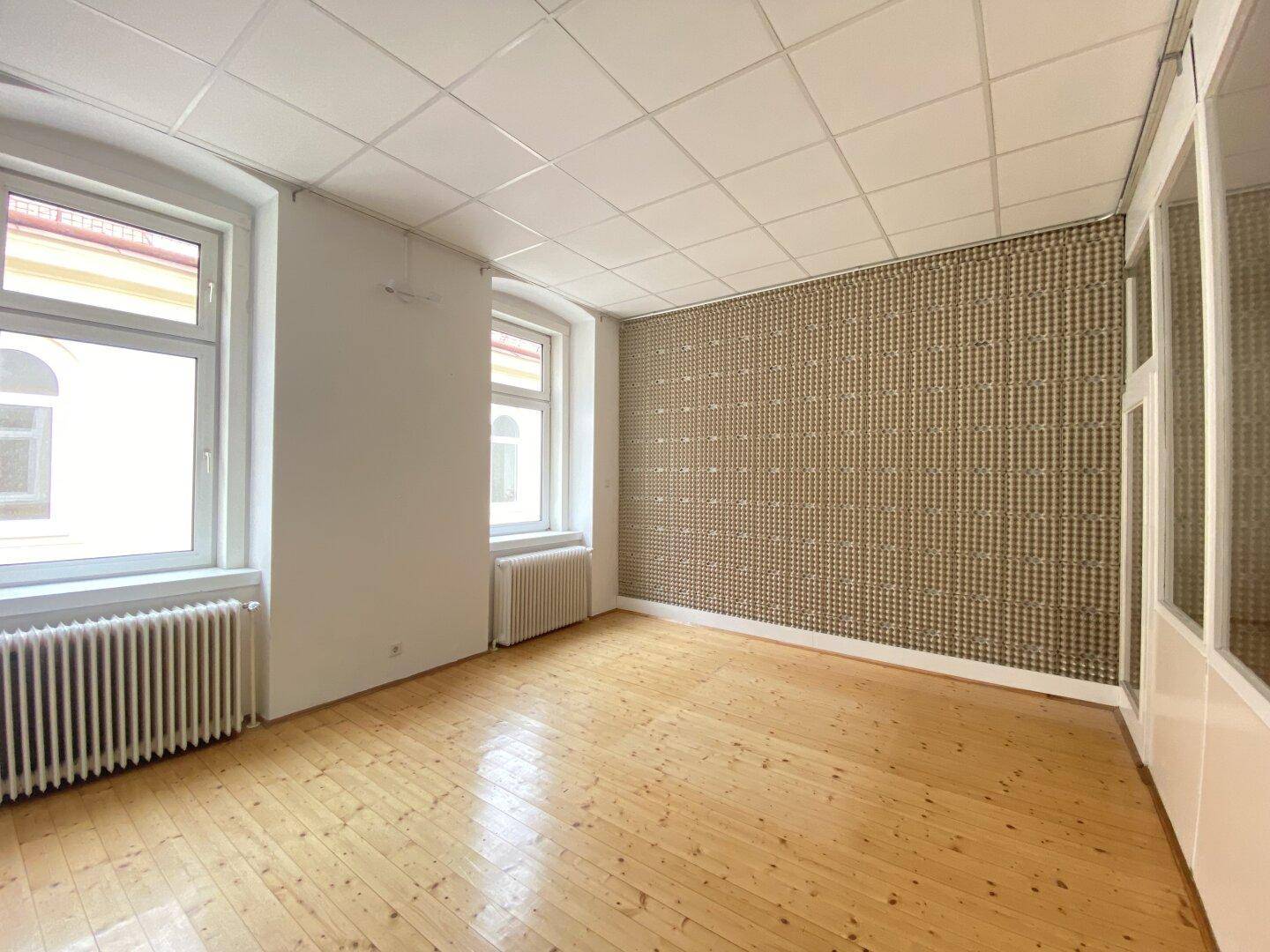 Büroraum mit Holzplankenboden, Rasterdecke und einer gemusterten Tapetenwand, hell durch zwei Fenster.
